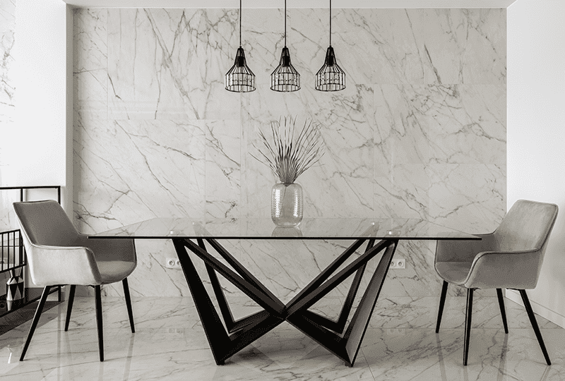 Modern dining area featuring a sleek tempered glass table with a unique base, surrounded by stylish chairs, complemented by a marble wall and pendant lighting, showcasing custom glass design elements.