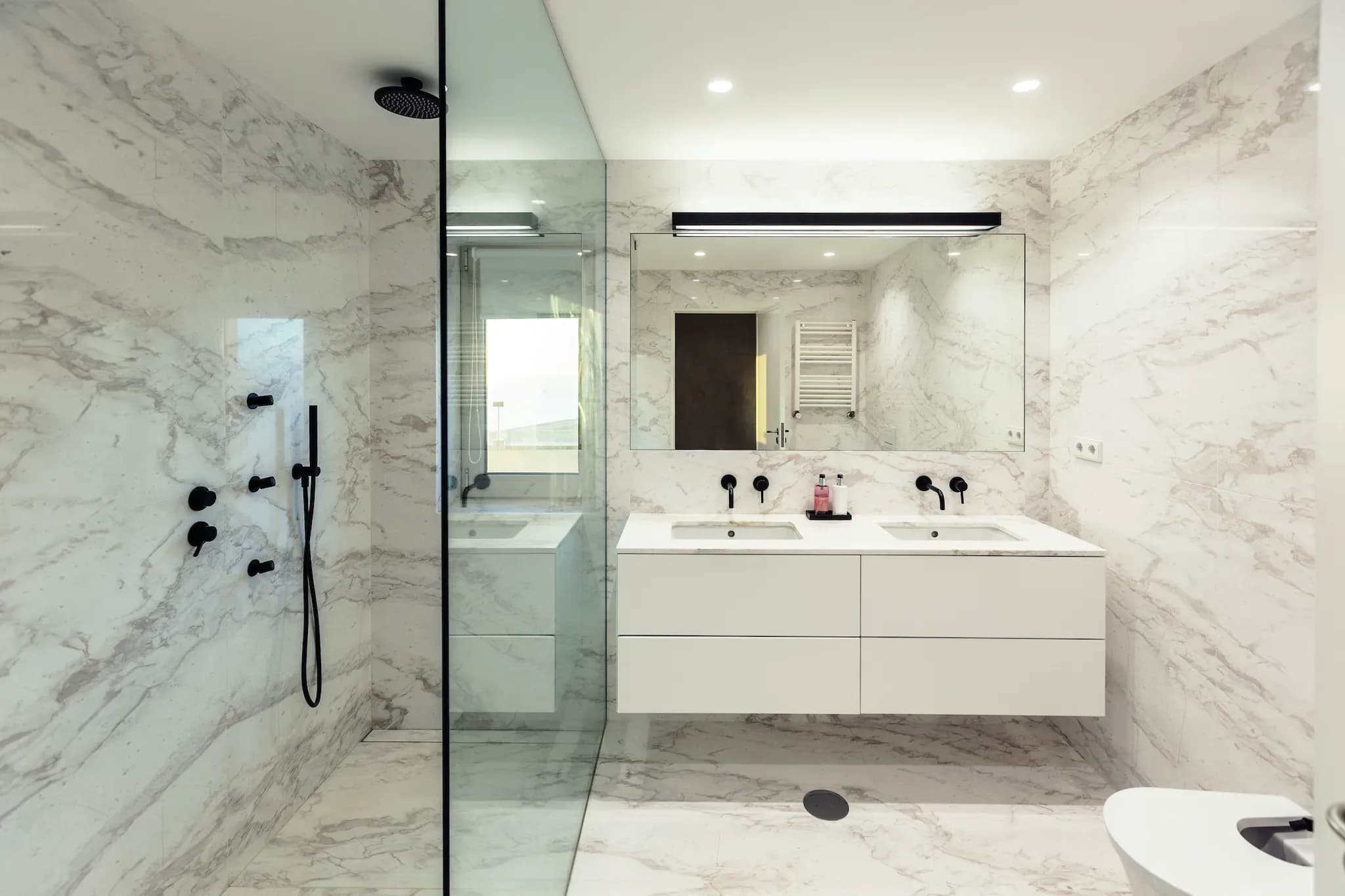 Bathroom setting with marble floor and walls. The focus is on a glass shower door seen from the side, with a rectangle mirror and sink in the background
