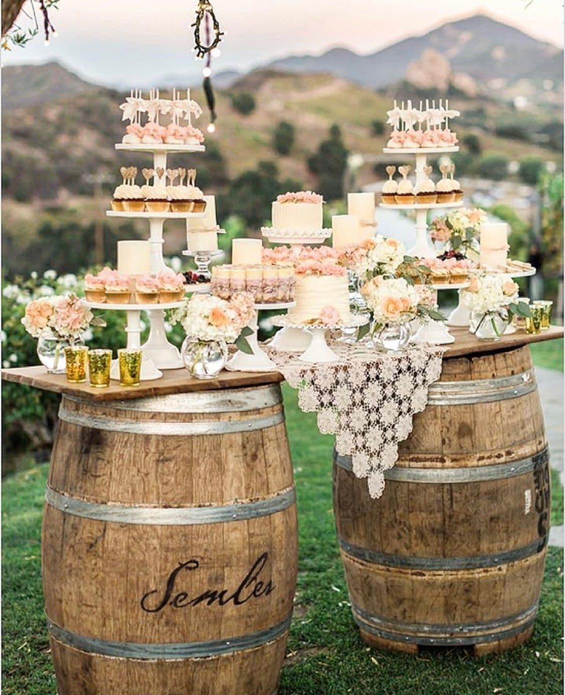 Elegant outdoor dessert setup featuring cakes, cupcakes, and floral arrangements on wooden barrels, showcasing custom glass serving platters and mirrors for a stylish event.