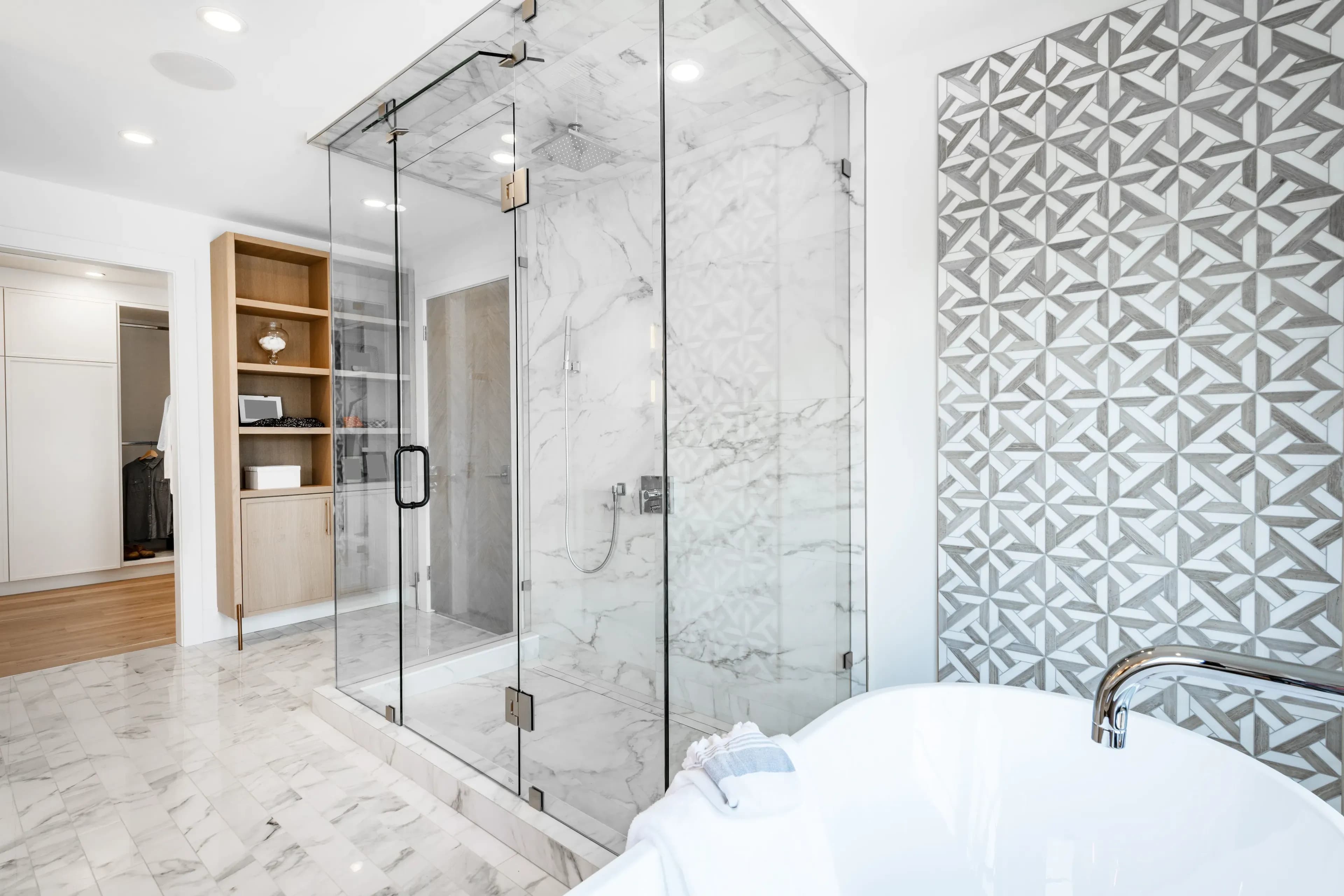 Grey bathroom, granite floor. Large shower, brushed chrome hardware, complex wall tile. White bathtub, wooden shelf. Hallway in background.