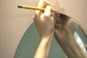 A hand holding a pencil marks a template on a beveled glass mirror