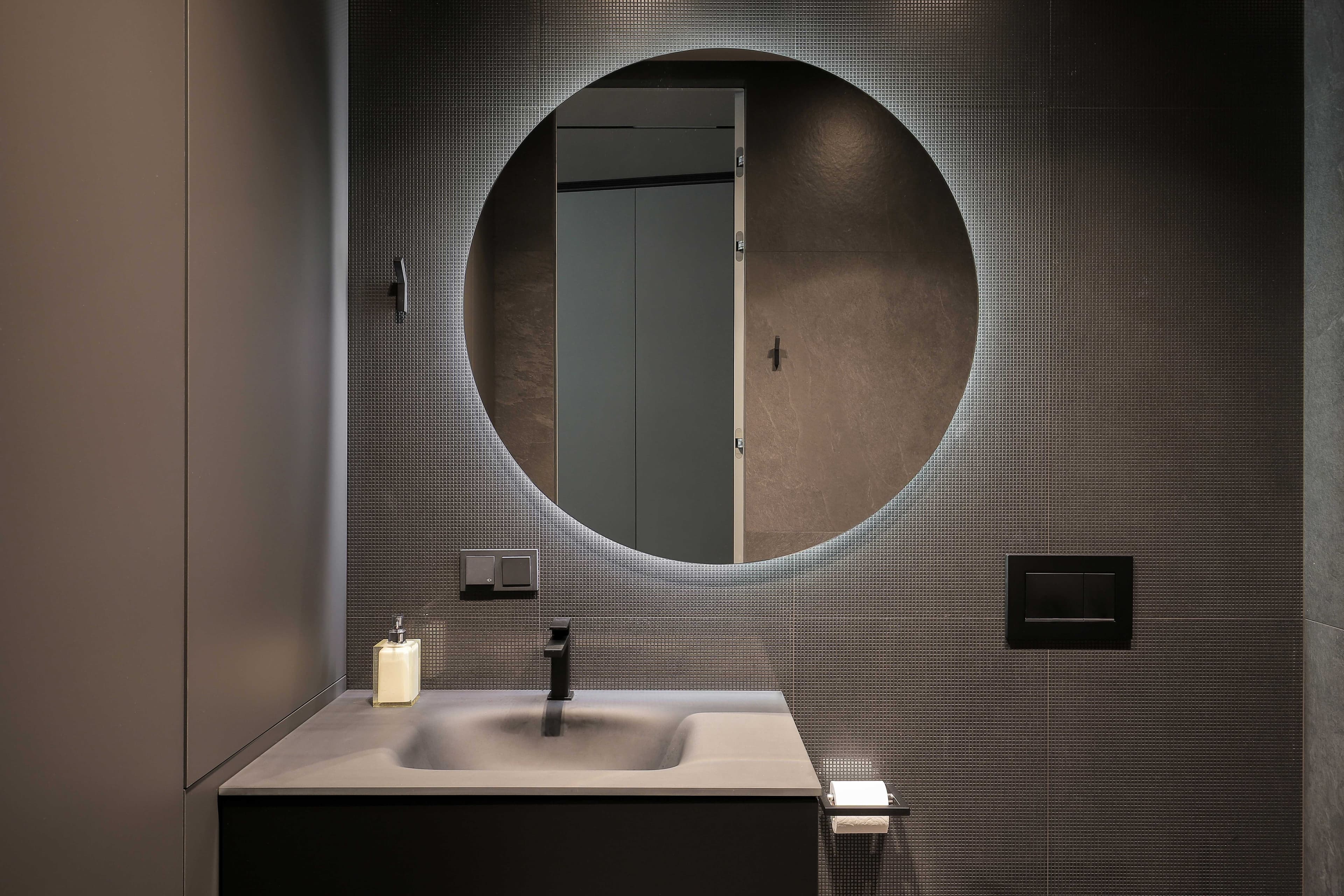 Modern bathroom with a circular illuminated mirror, sleek black sink, and stylish fixtures, showcasing custom glass design elements relevant for shower doors, tempered glass, and beveled glass edges.