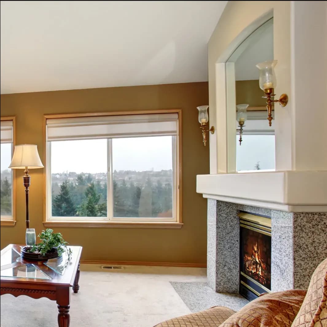 A cozy living room featuring a decorative mirror above a fireplace, a stylish table with a plant, and large windows allowing natural light, showcasing custom glass solutions like mirrors and tempered glass accents.