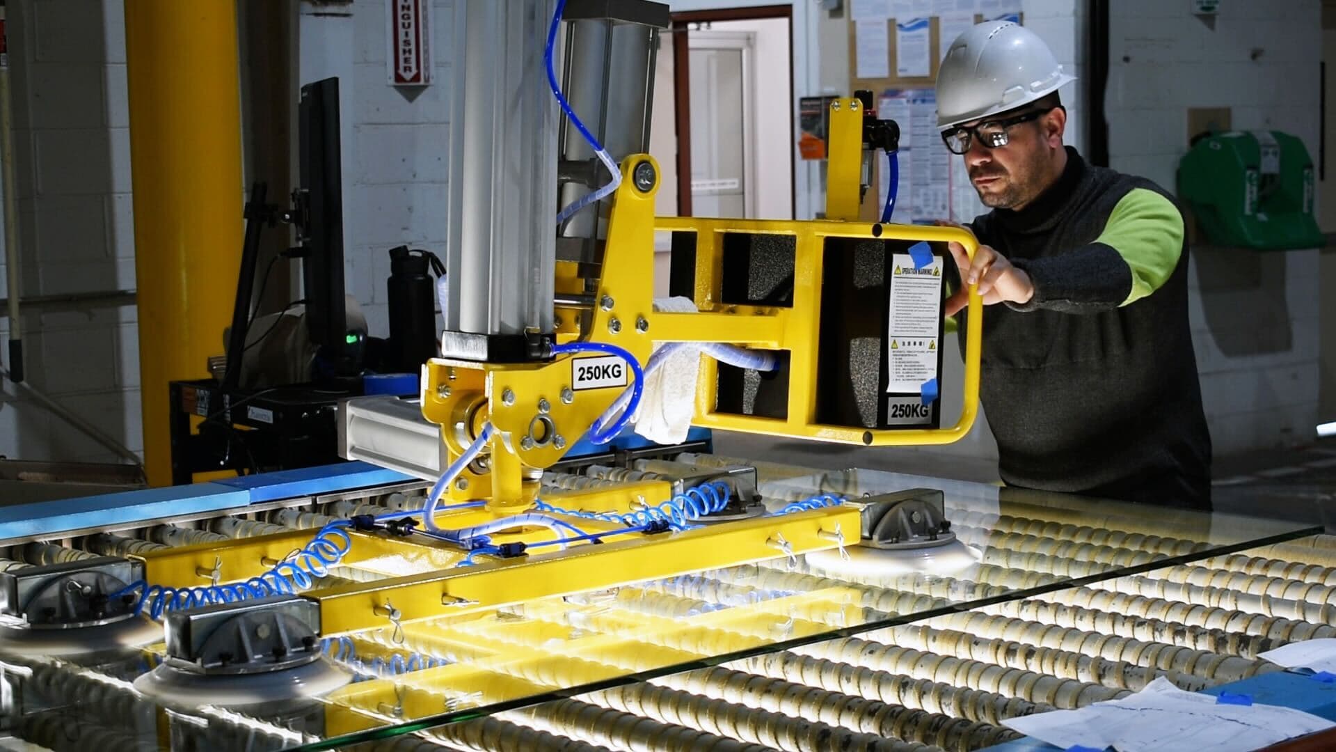 Worker operating glass handling equipment for custom glass products, including shower doors and table tops, highlighting the use of tempered glass and beveled glass edge techniques in a manufacturing environment.