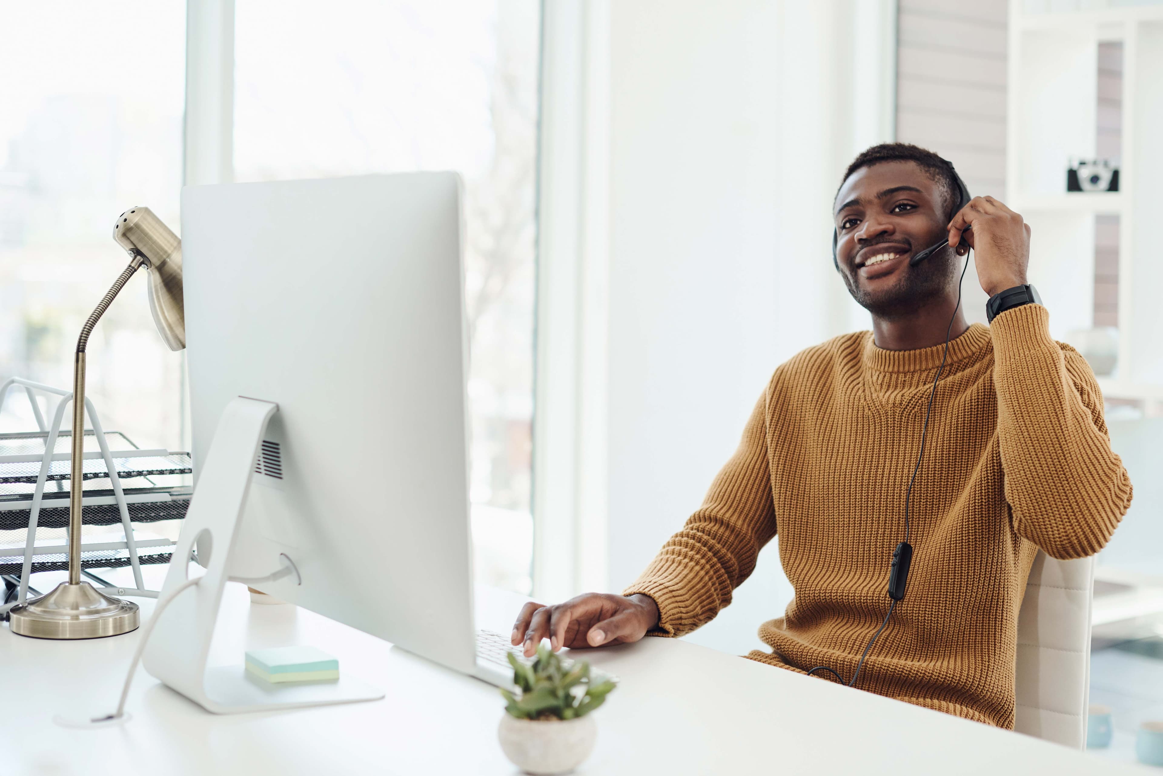 A smiling man in a mustard sweater sits at a desk with a sleek computer, engaged in a conversation using a headset, representing a modern workspace for customer service.