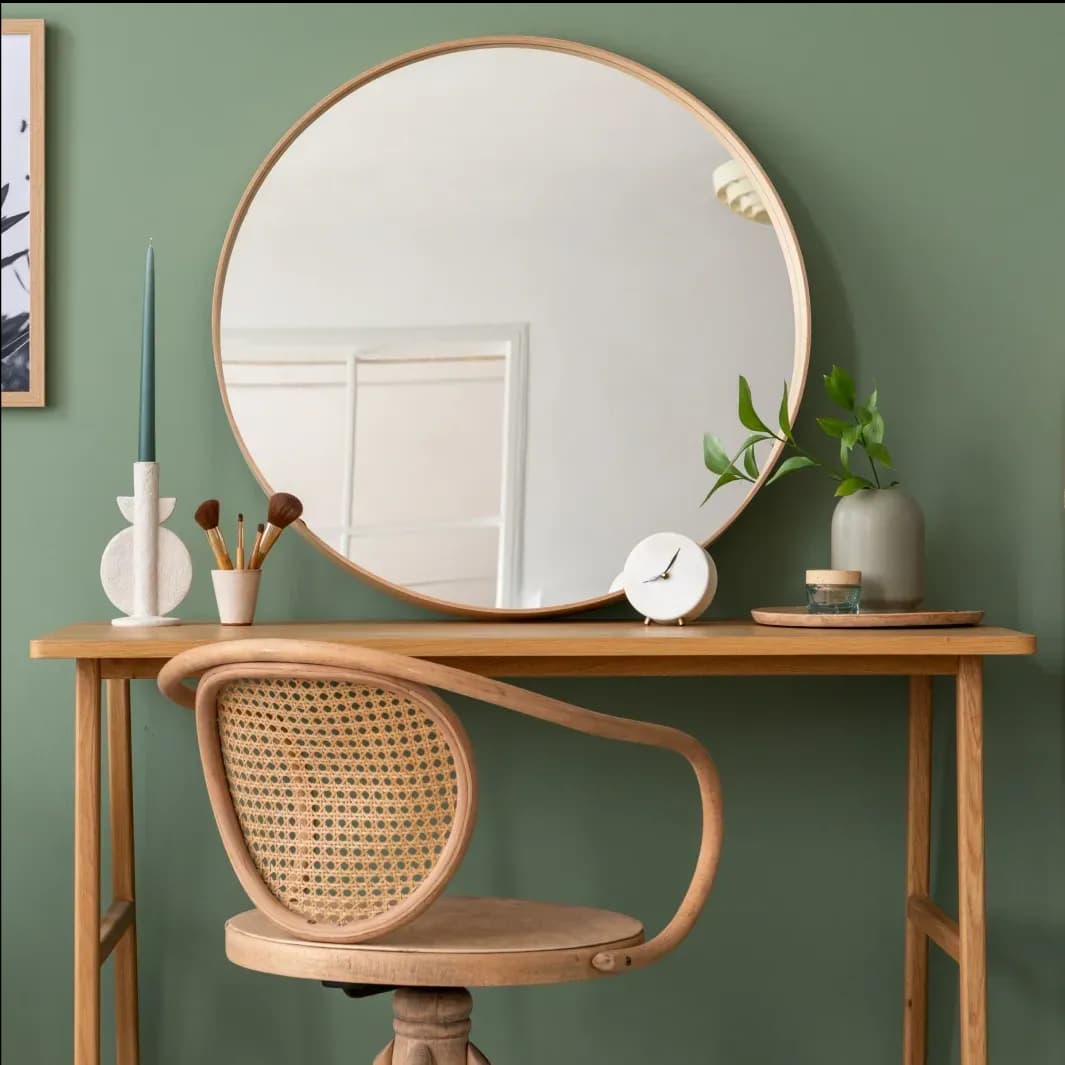 Modern workspace featuring a round custom mirror with a wooden frame, a light green wall, wooden table, and decorative elements, highlighting the elegance of glass design in interiors.