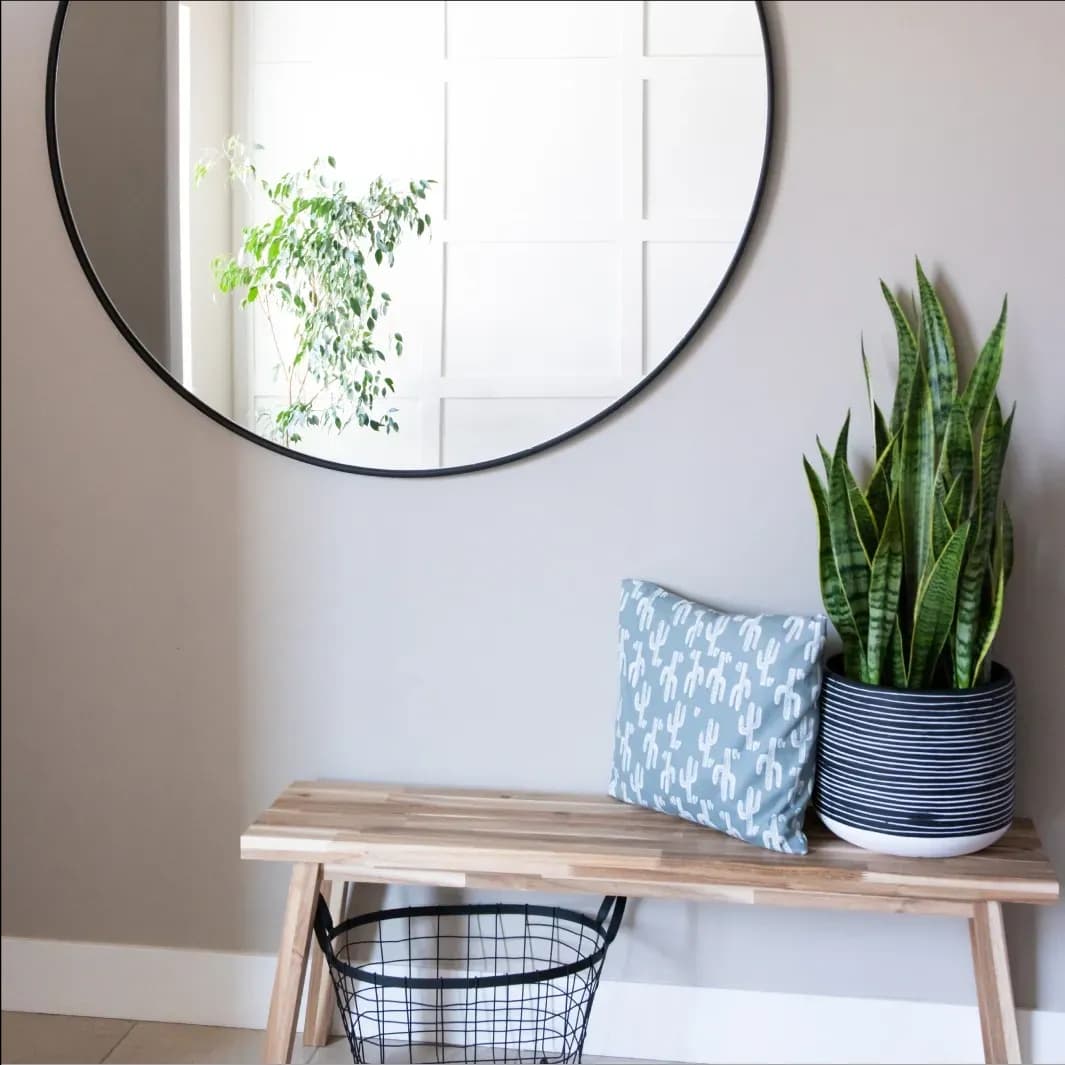 Modern interior featuring a round mirror with a black frame, a minimalist wooden bench, decorative pillow, and indoor plants, highlighting custom glass design for mirrors and home decor.