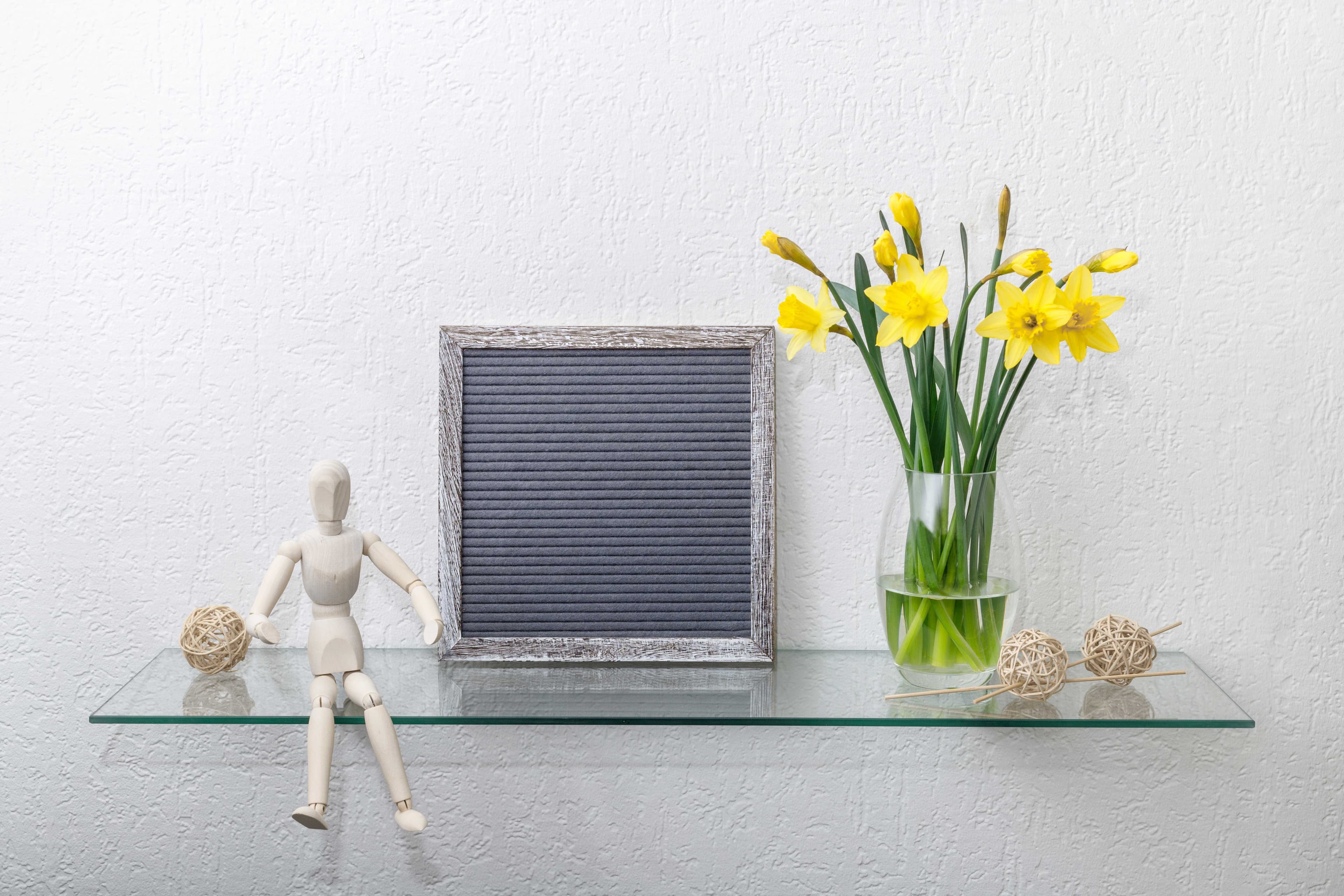Glass tabletop displaying a wooden mannequin, decorative string balls, a framed notice board, and a vase of yellow flowers, highlighting custom glass options and contemporary decor.