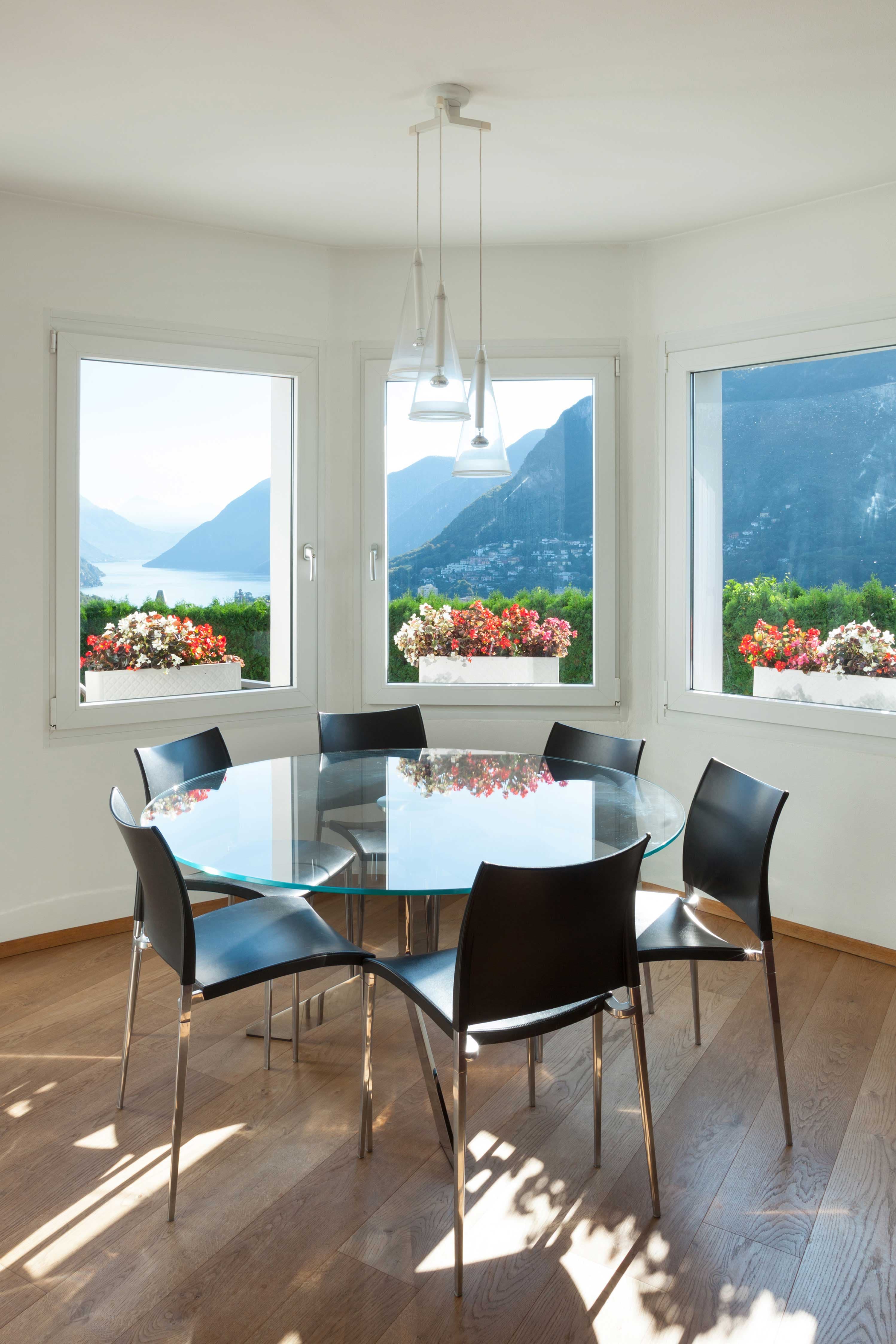 Modern dining area featuring a round glass table with a beveled glass edge, black chairs, and large windows showcasing stunning mountain views. Ideal setting for showcasing custom glass designs.