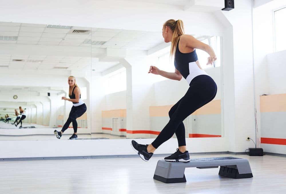 A woman exercising on a step platform in a bright gym with large mirrors
