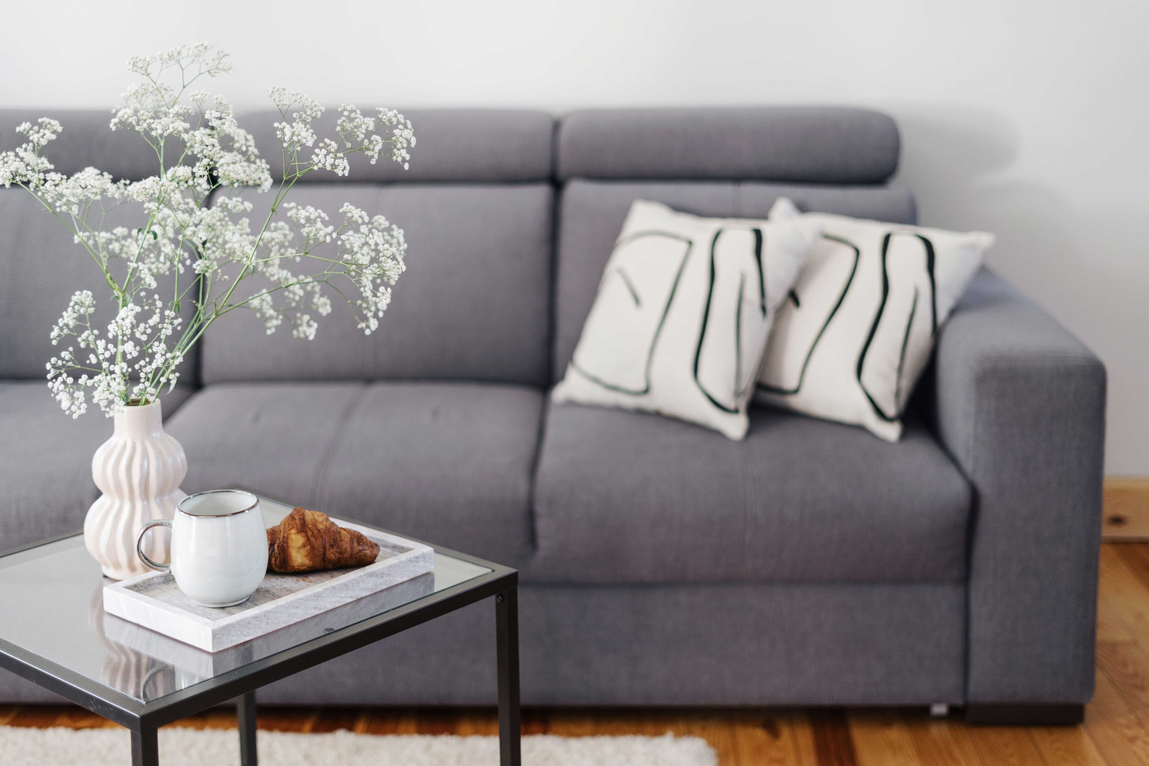 Cozy living room scene featuring a gray sofa, decorative pillows, a coffee table with a custom glass top, a vase with flowers, a mug, and a croissant, highlighting modern design elements.