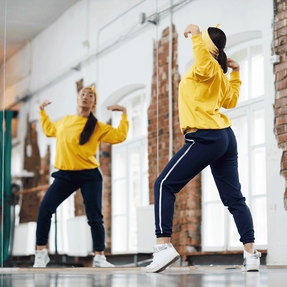 A dancer in a yellow outfit practices in front of a mirror, showcasing the studio's spacious design ideal for showcasing custom glass features like mirrors and shower doors.