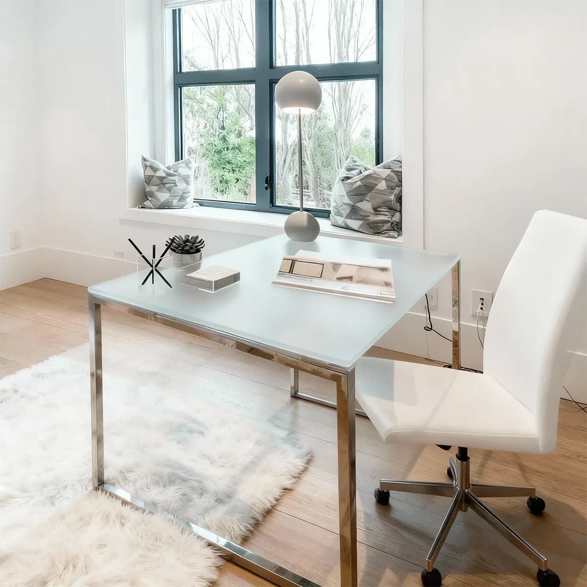 A bright home office featuring a modern desk with a translucent, frosted glass top and a polished chrome frame, paired with a white office chair.