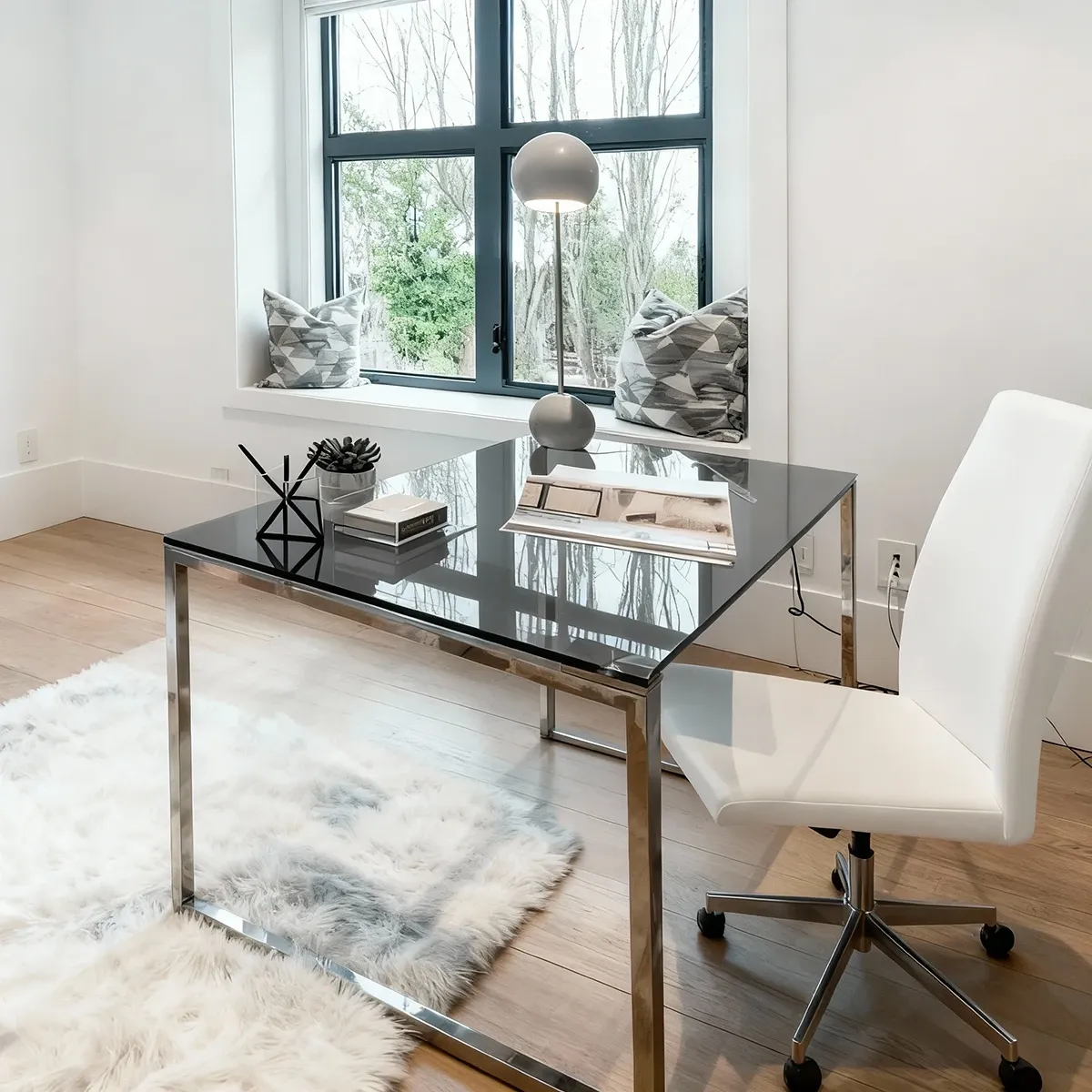A bright home office with a sleek, modern desk featuring a reflective, grey-tinted glass top and a polished chrome frame, paired with a white office chair.