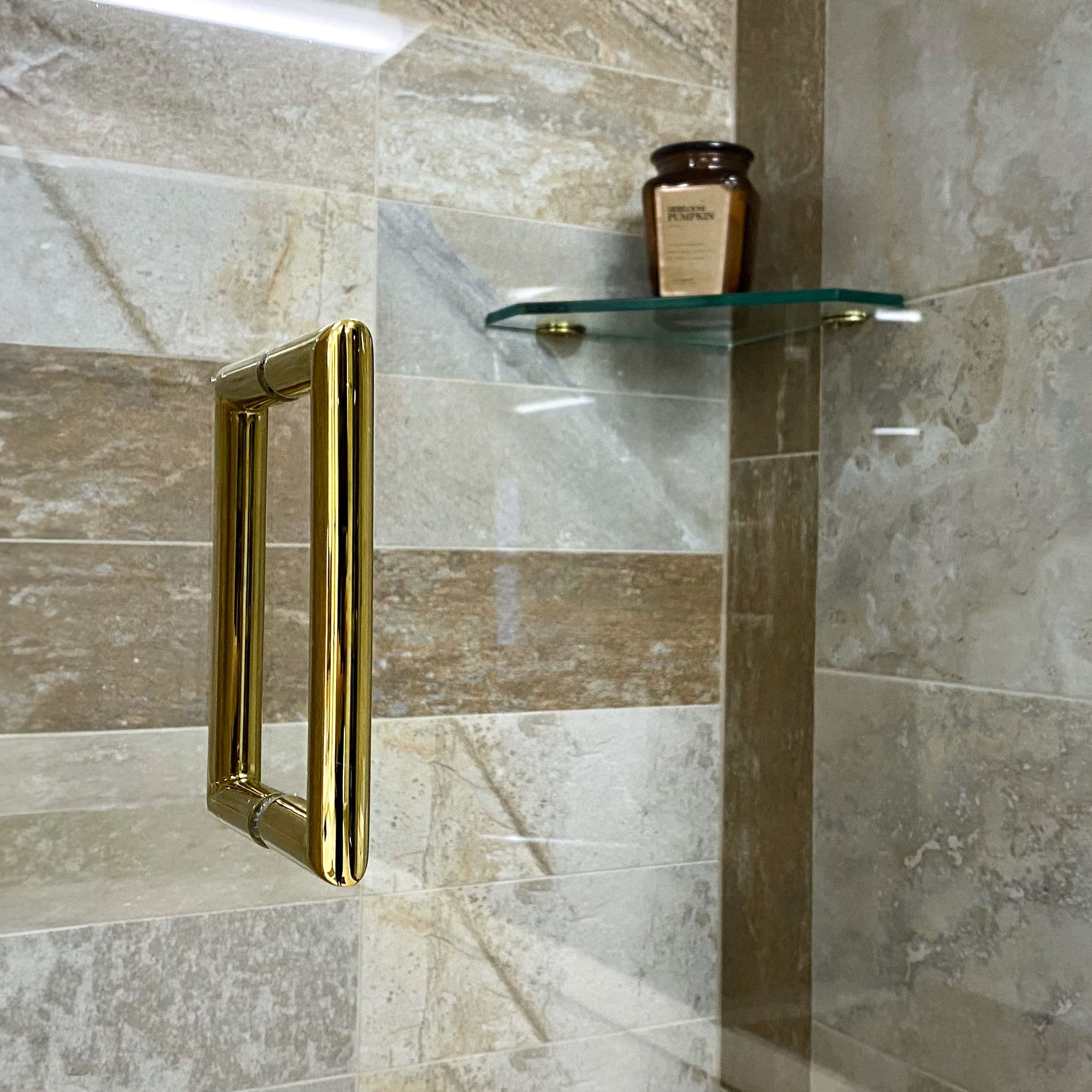 Close-up of a gold shower door handle alongside a glass shelf, showcasing custom glass designs and tempered glass finishes, ideal for modern bathrooms.