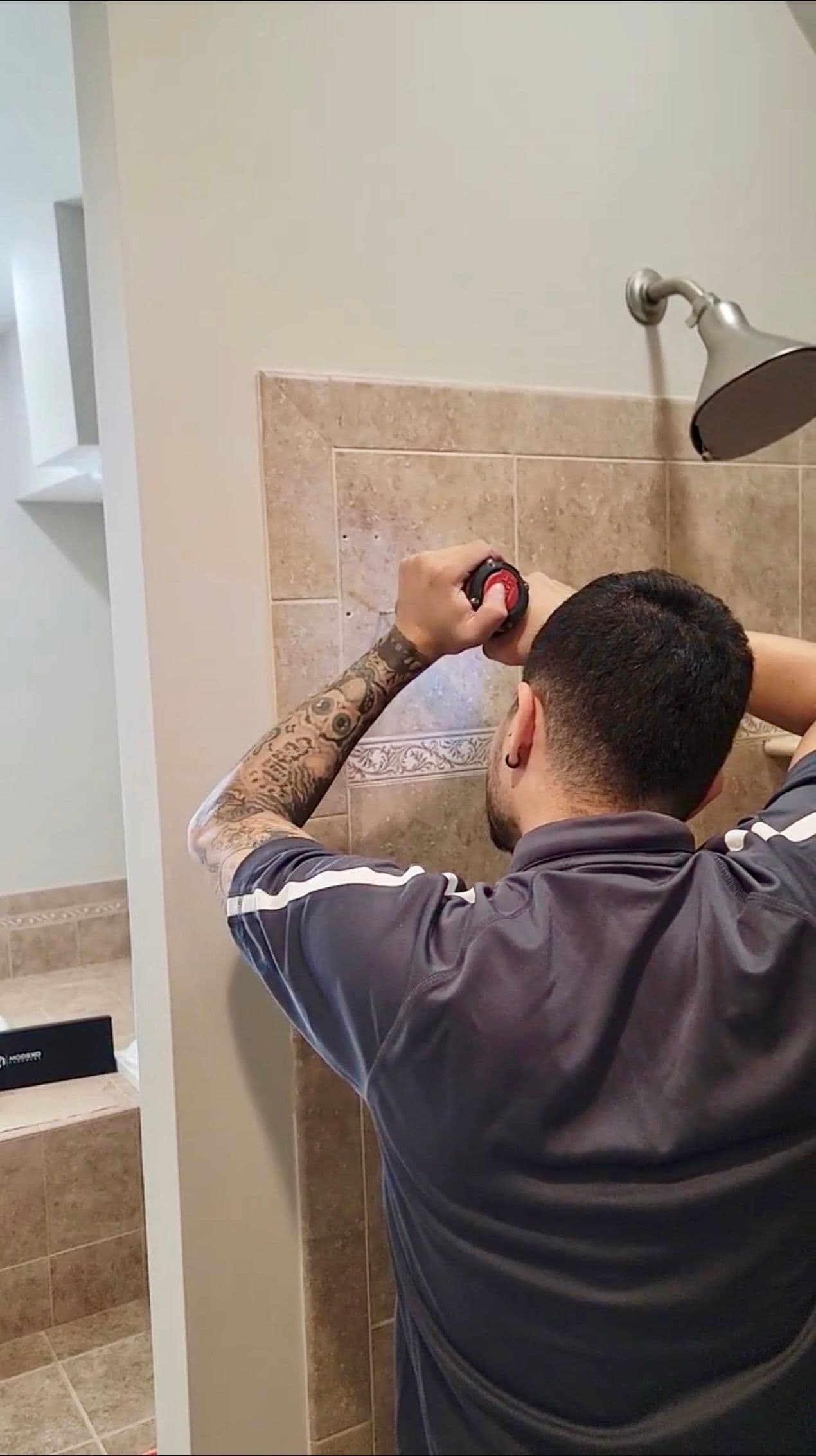 A technician with a tattooed arm uses a device to mark a spot on a tiled shower wall, preparing to install a fixture.