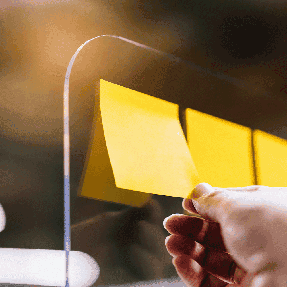 Hand placing a yellow sticky note on a custom glass surface, showcasing the durability of tempered glass for shower doors, table tops, and mirrors with a beveled glass edge.