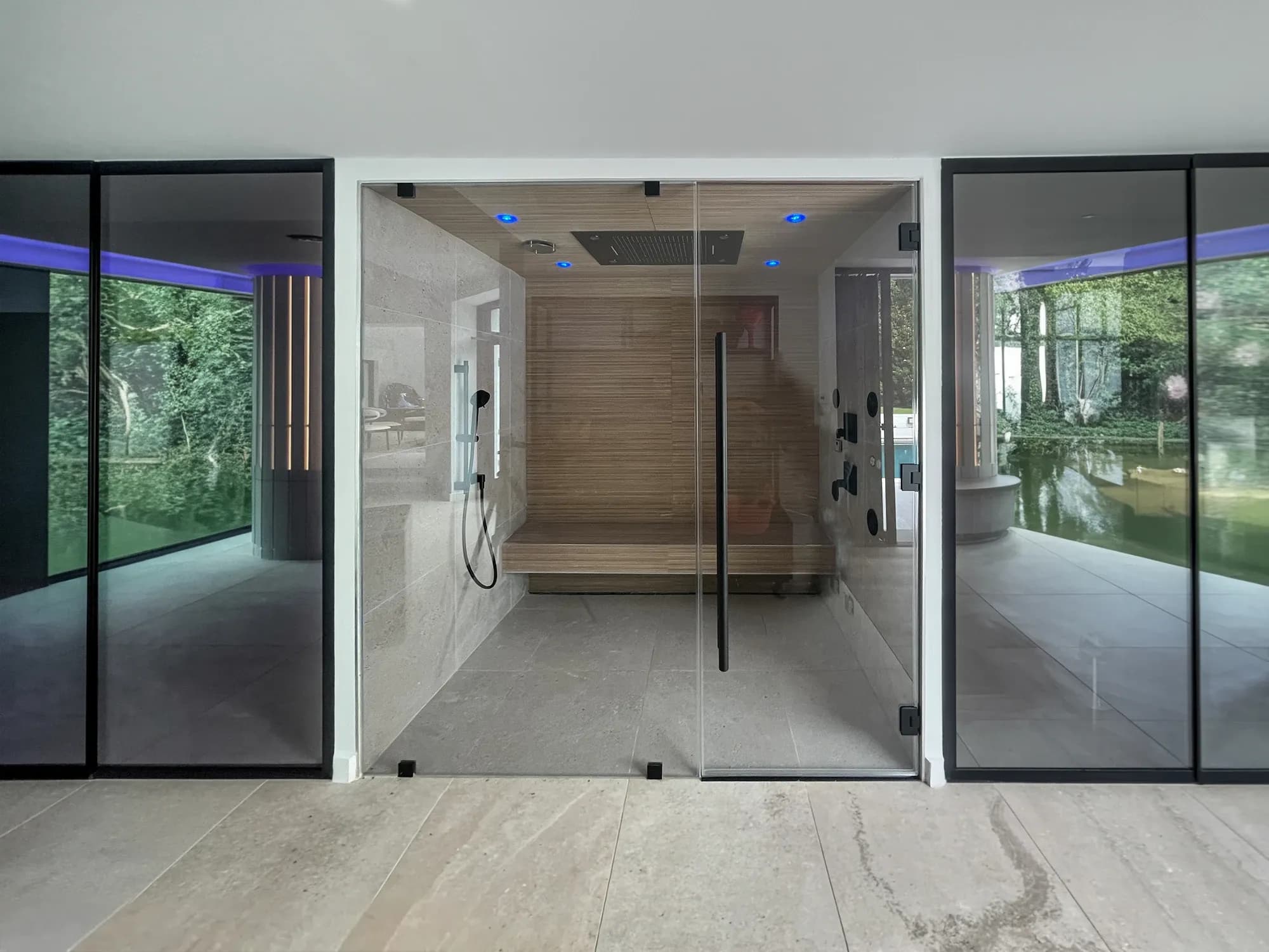 A modern built-in steam room and shower with a black-framed glass front, wood bench, and blue ceiling mood lighting.