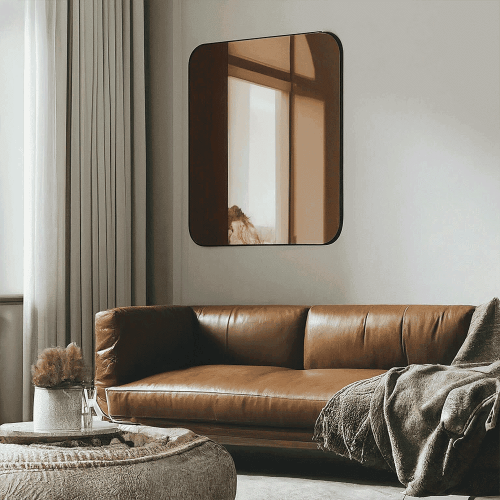 Custom glass mirror with beveled edges mounted on a wall above a stylish brown leather couch in a modern living room setting, showcasing tempered glass design elements.