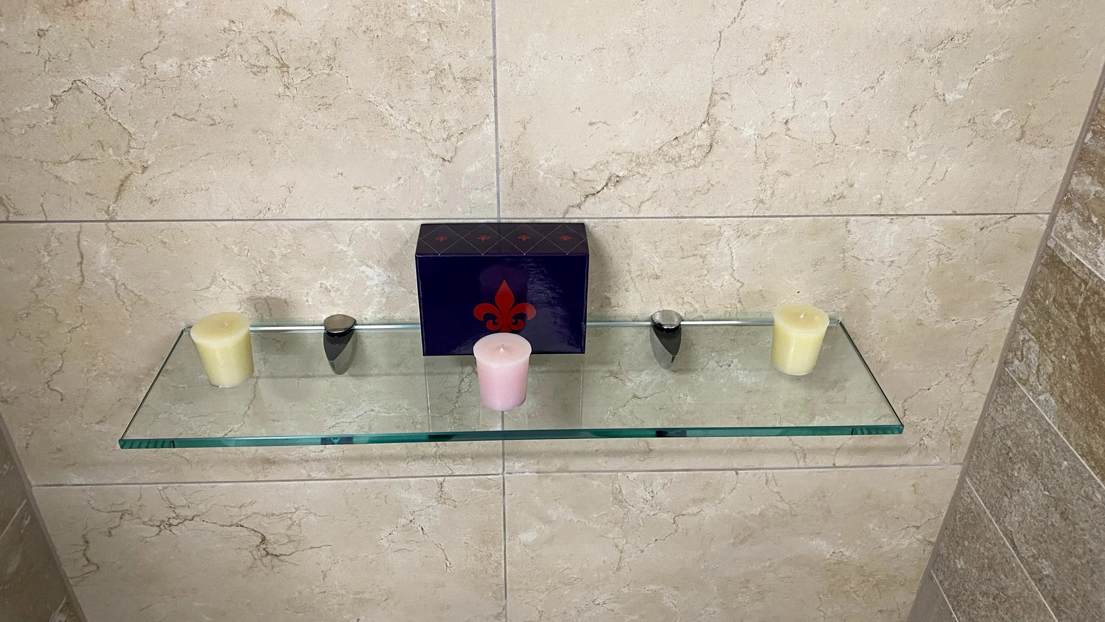 A rectangular glass shelf with chrome brackets mounted on a tiled bathroom wall, holding decorative candles and a small box.