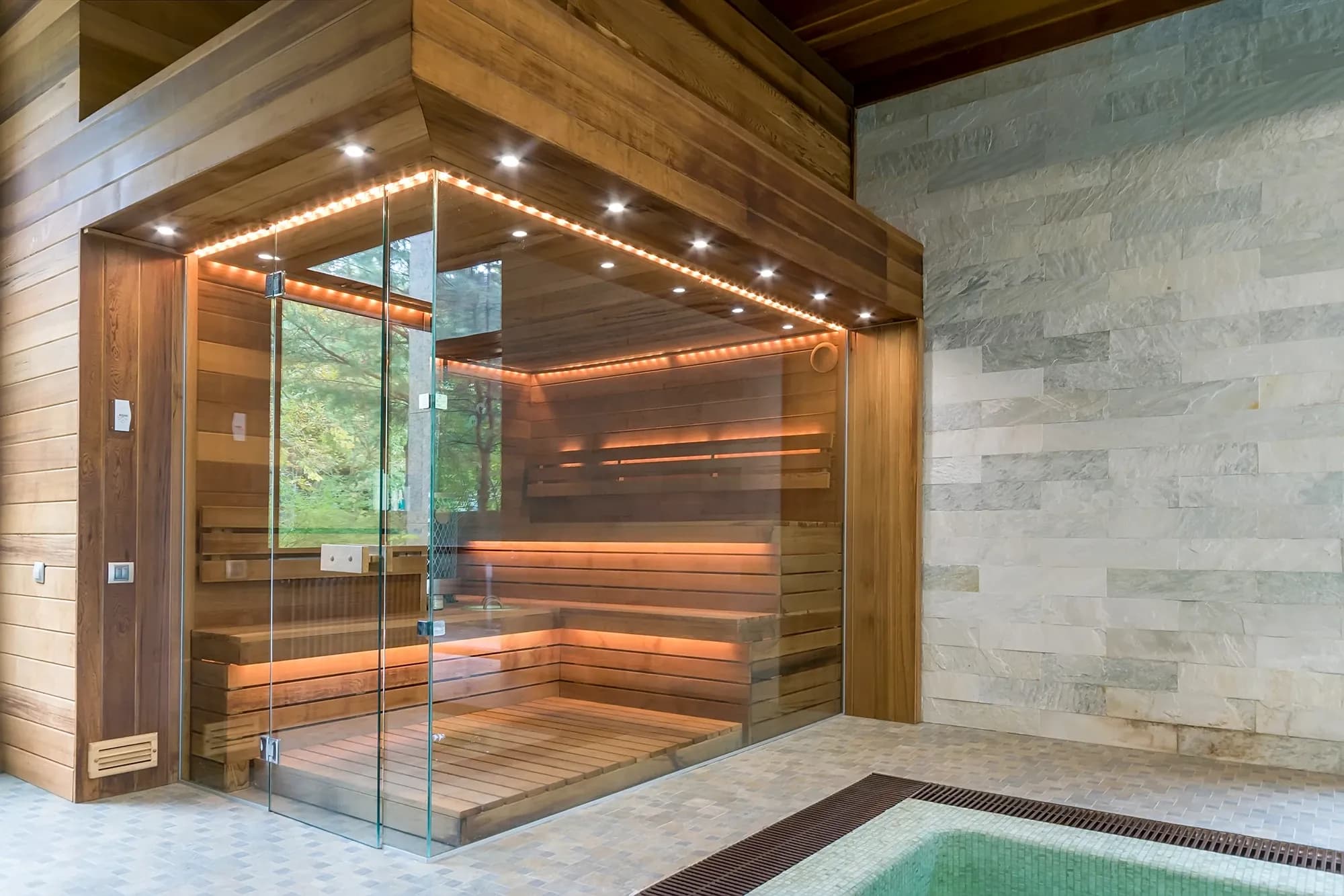 A modern custom-built sauna with a glass front, wood interior, and warm LED accent lighting next to a stone wall and plunge pool.
