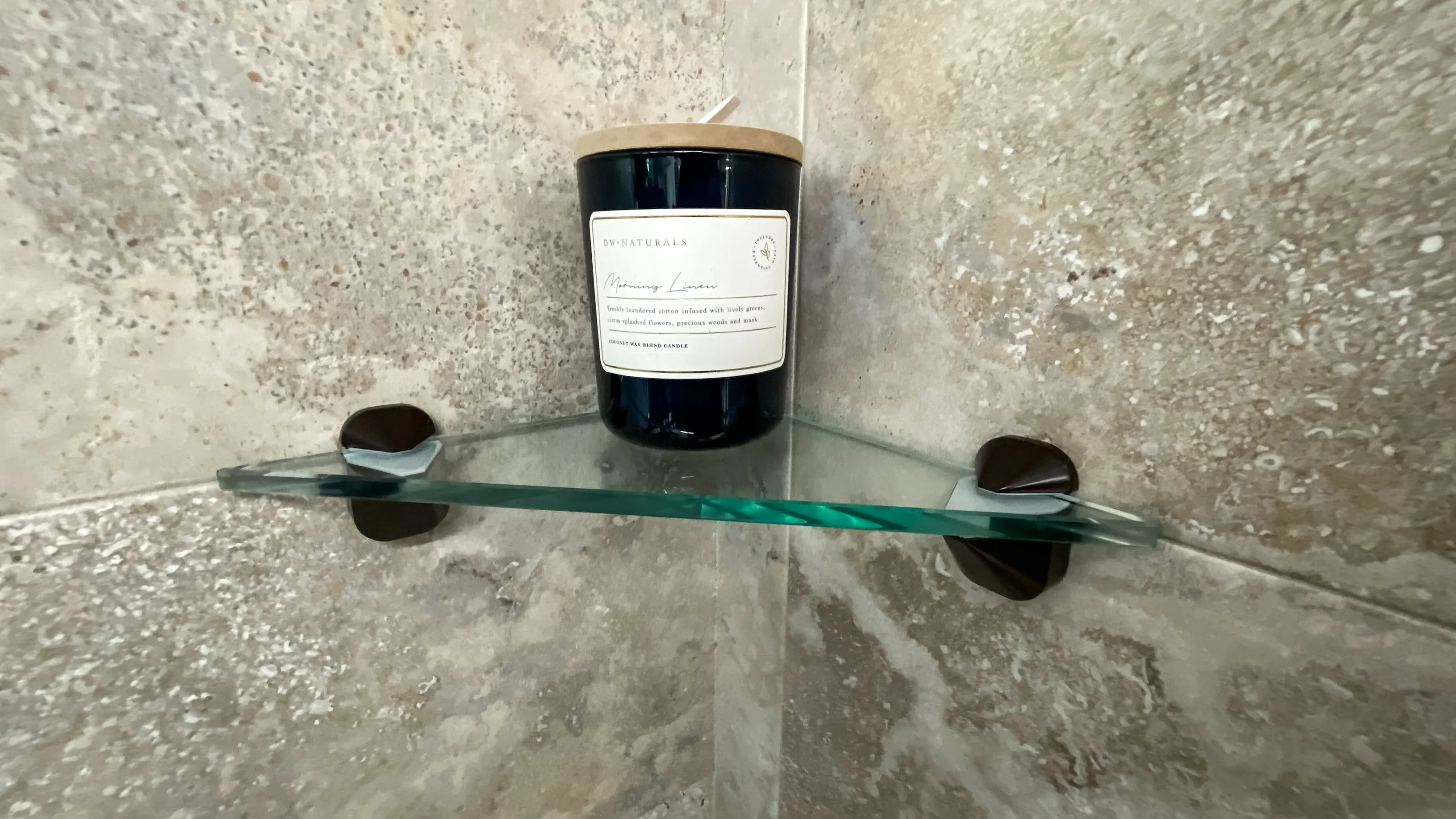A curved corner glass shelf with oil-rubbed bronze brackets, installed in a tiled shower and holding a large candle.