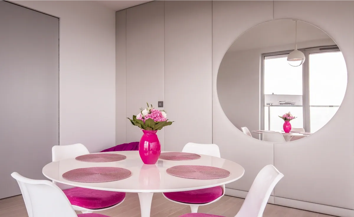 white dining room with pink chair cushions, place mats, and vase. A large round mirror hangs on the back wall