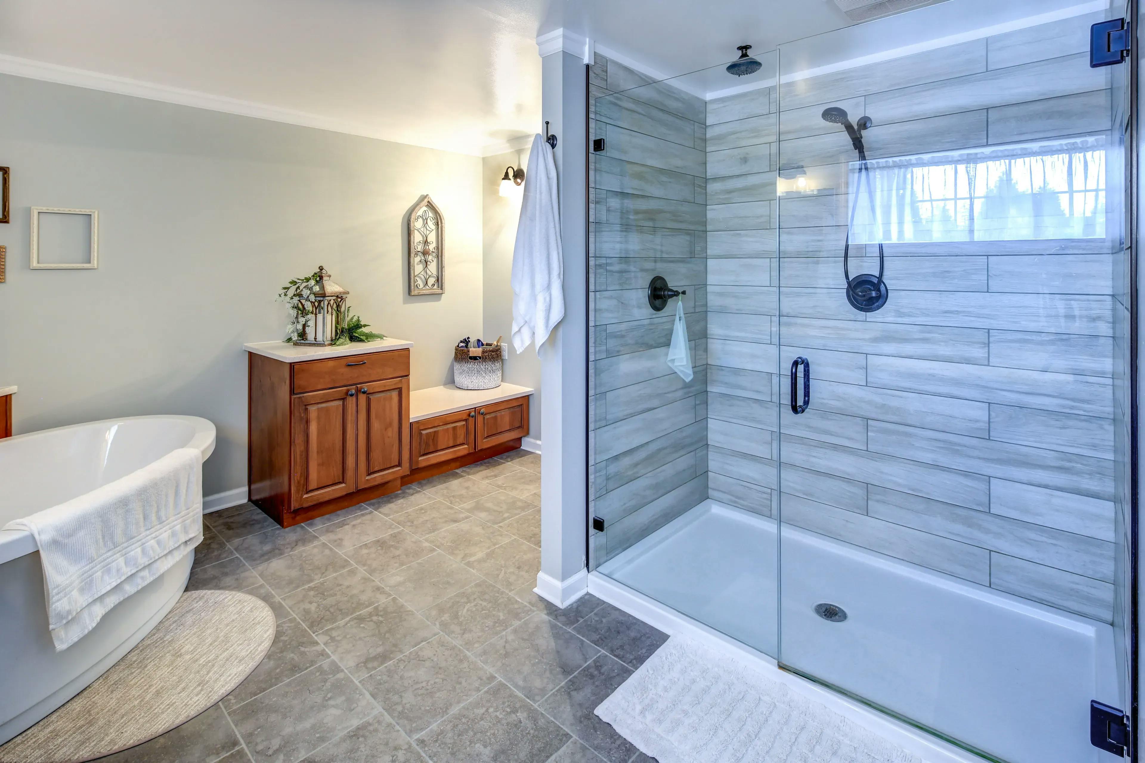 Grey tile floor, beige tile walled shower with black hardware, white shower rug, bathtub, white granite sink with wood shelving in a grey bathroom.