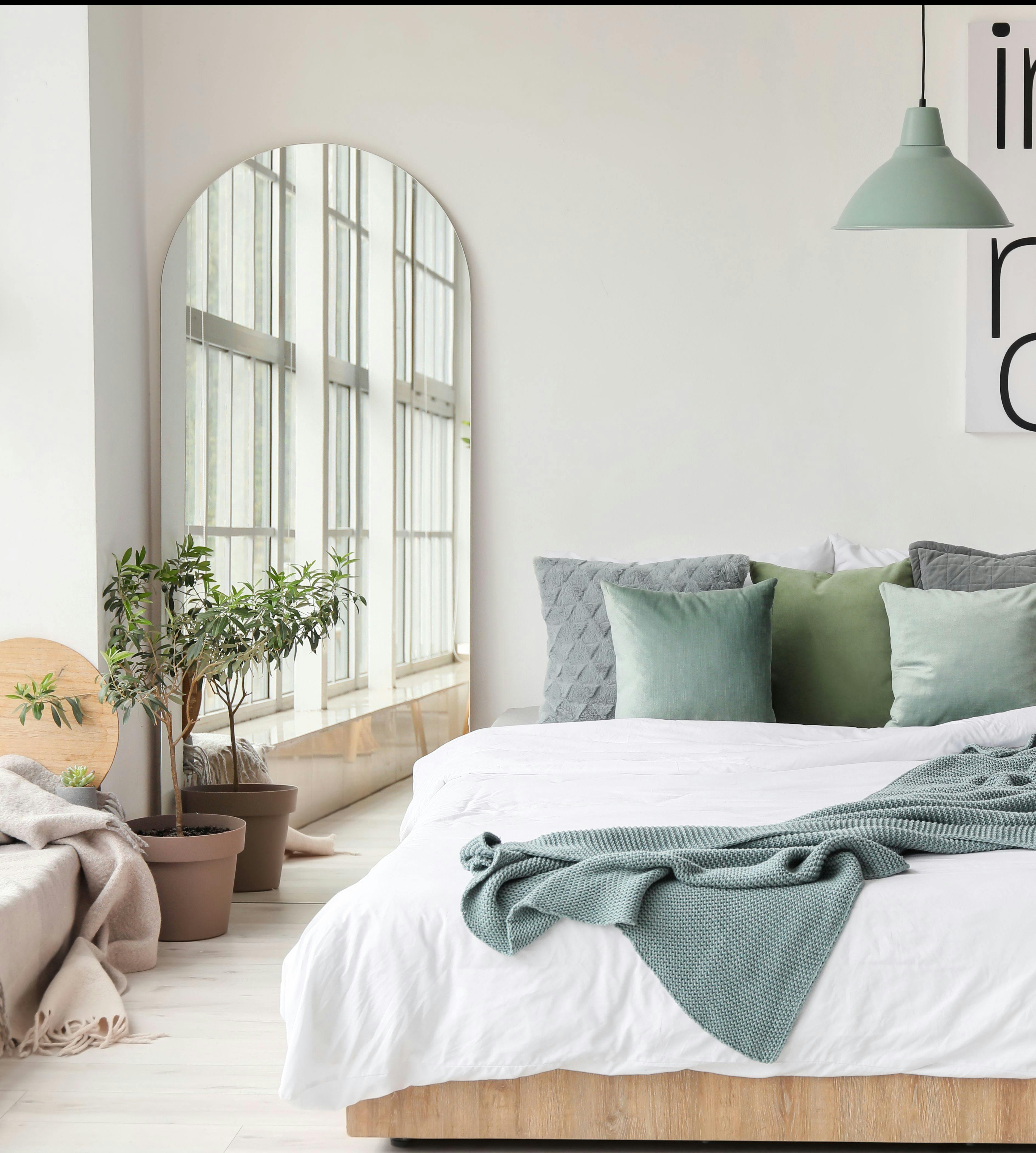 A serene white bedroom featuring a bed with green pillows and white bedding, adorned with a green throw blanket. A curved oval mirror leans against the wall, accompanied by a vibrant potted plant in a planter placed at its base. Light wood floors complete
