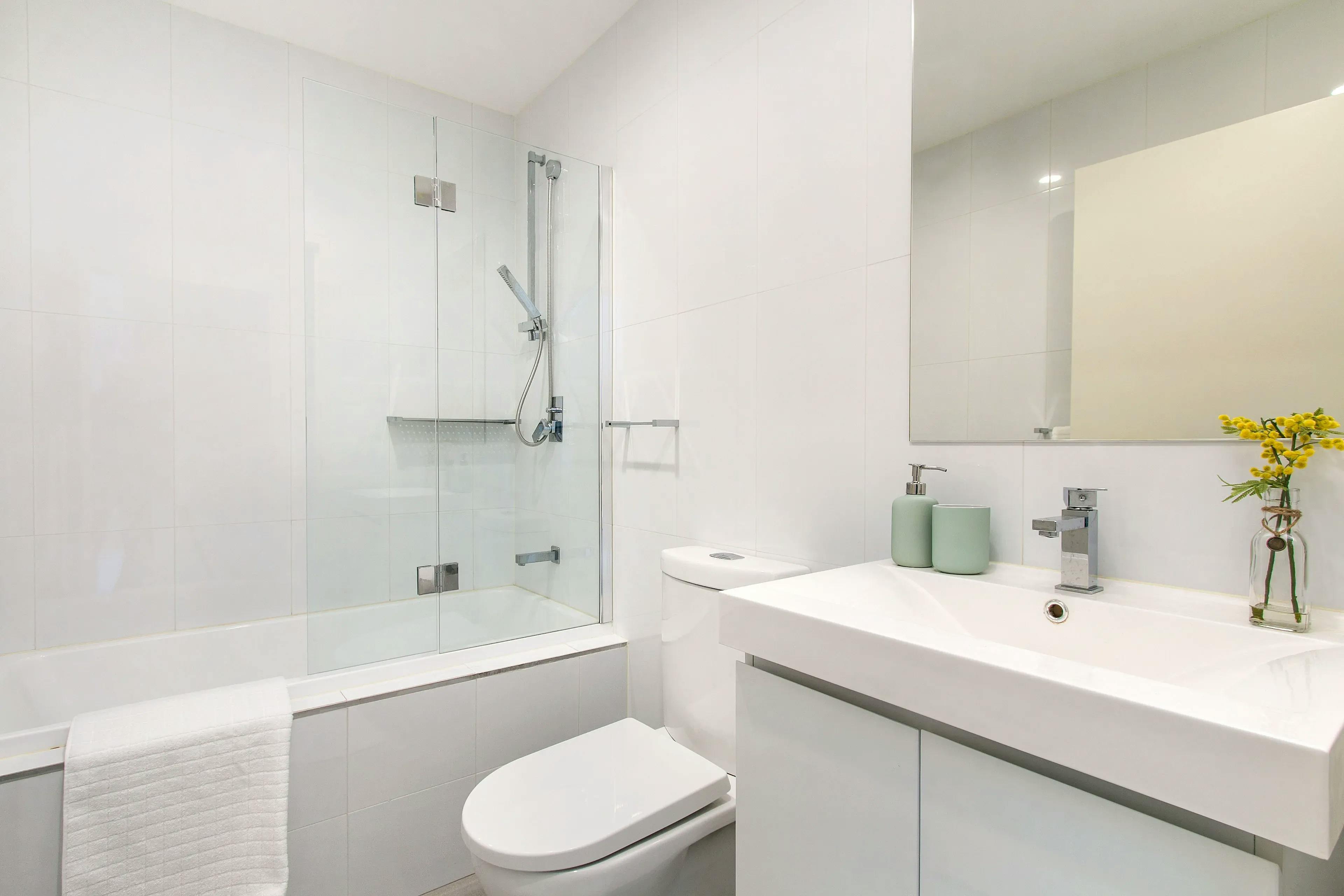 Bathroom setting with all-white walls, a bathtub featuring a glass shower panel, and a white sink accompanied by a rectangular mirror