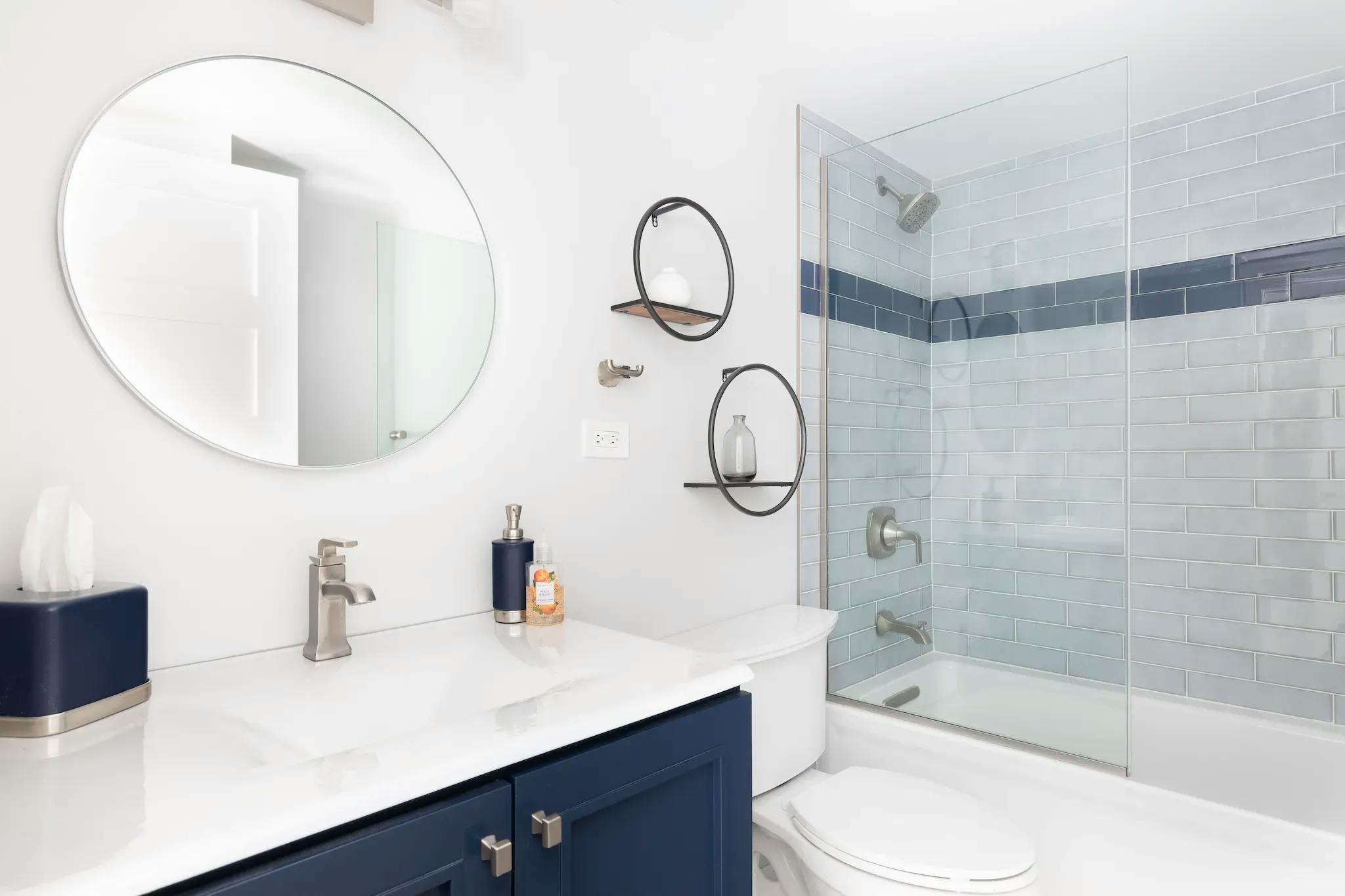 White bathroom, sliding shower door, blue strip on white tile, chrome hardware. Round mirror above white granite sink, black shelving, toilet next to sink.