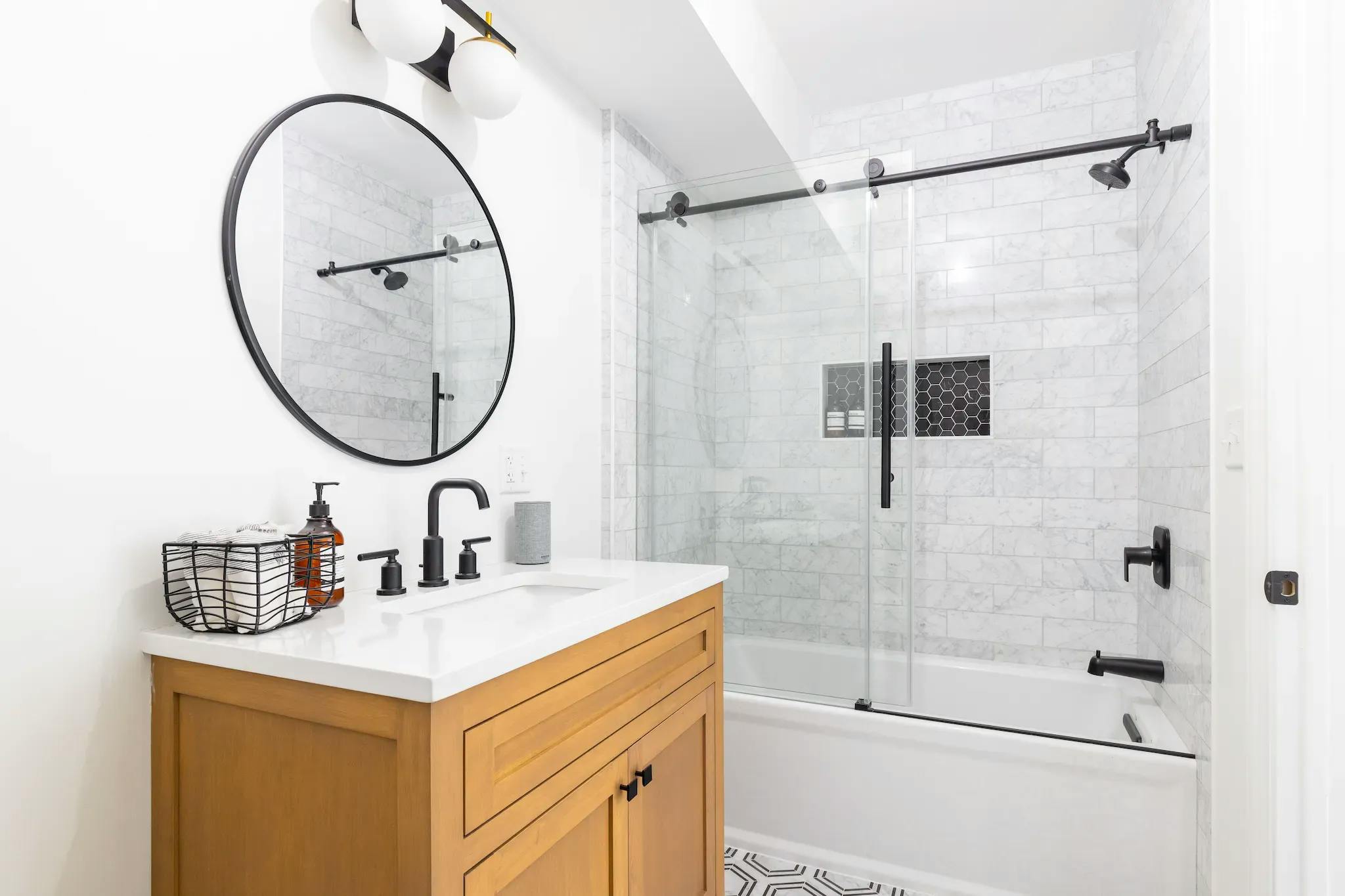 White bathtub, black hardware. Round mirror over white granite sink with soap and basket. Wooden shelving under the sink.