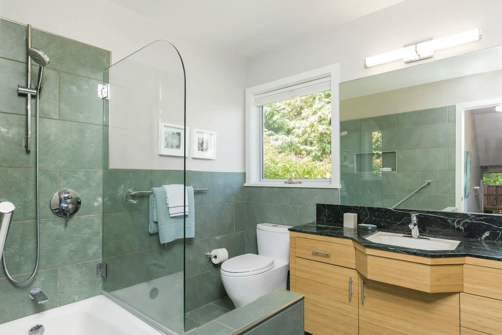 Green-tiled bathroom setting with a rectangle mirror above the vanity. The focus is on the glass panel over the bathtub