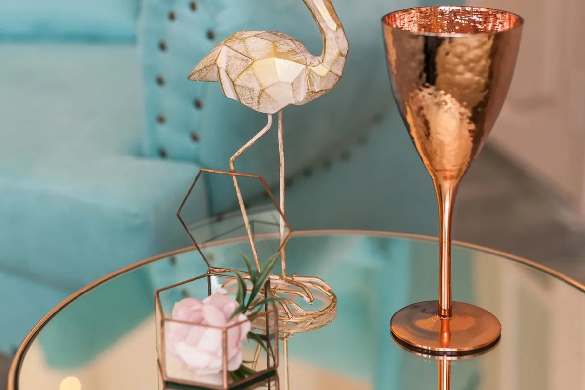 a close up of an end table with mirror top. The end table rame is brass, a rose gold candle holder, flamingo, and drink glass sit on top. 