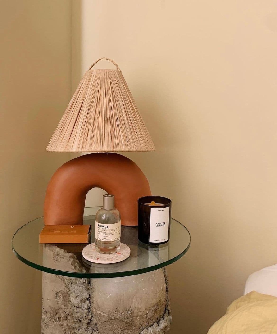 A modern side table featuring a custom glass top, showcasing decorative items like a candle and a perfume bottle, with a unique lamp made of ceramic and a textured concrete base.