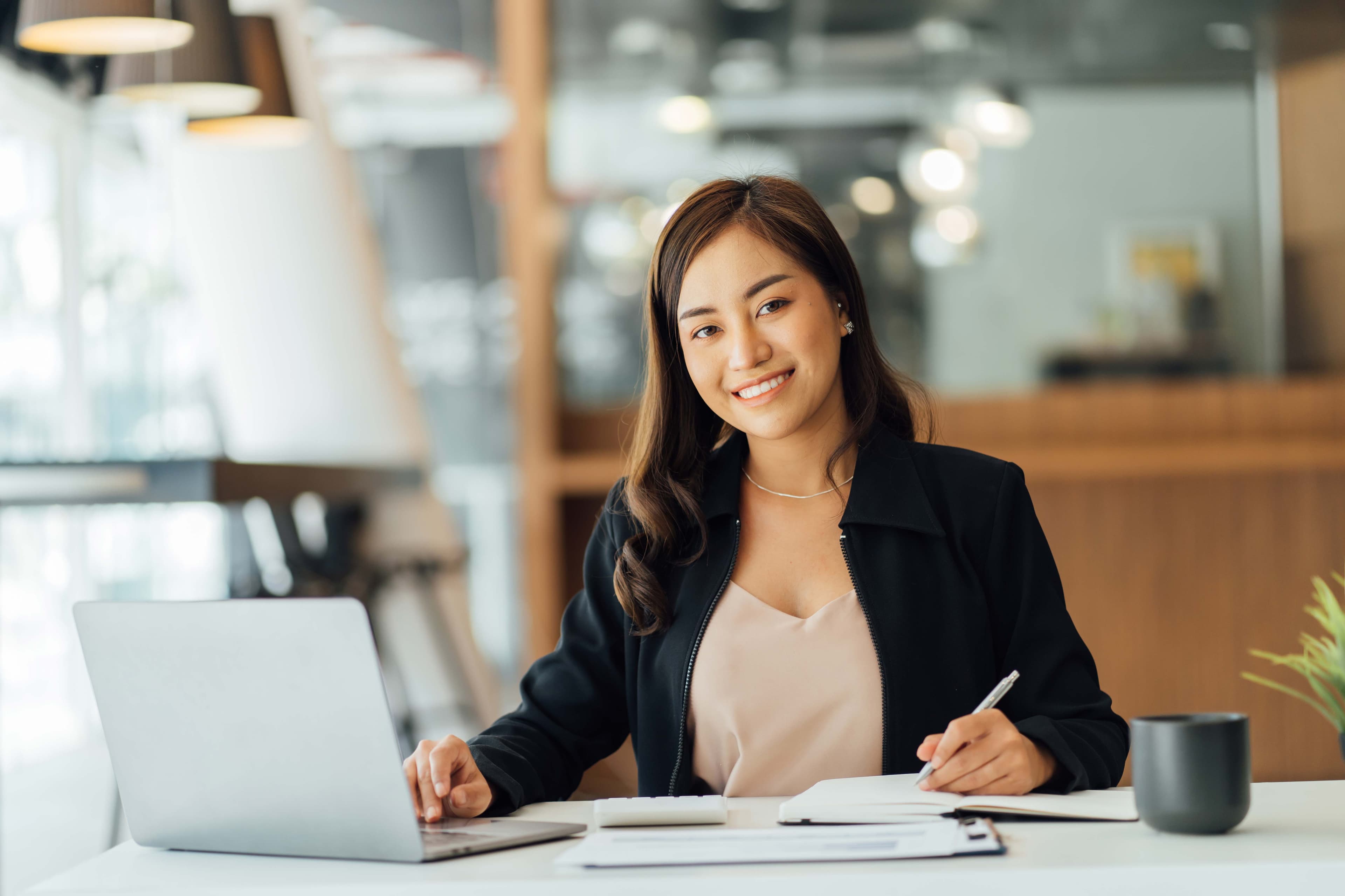 Smiling woman at a desk working on a laptop, with notepad and coffee cup, representing a modern office environment.