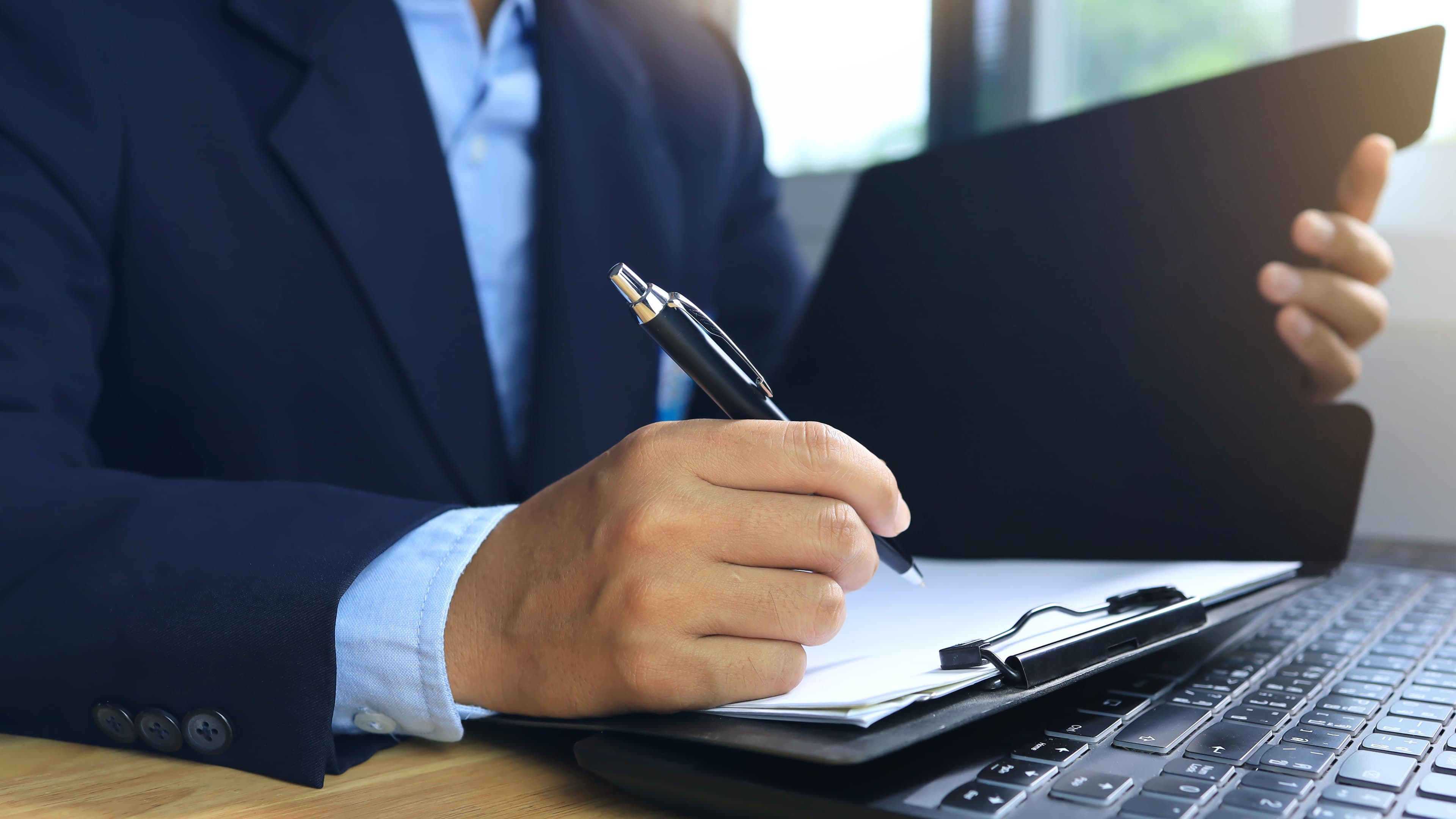 Business professional taking notes with a pen, surrounded by paperwork and a laptop, emphasizing custom glass solutions like shower doors, table tops, mirrors, tempered glass, and beveled glass edges.