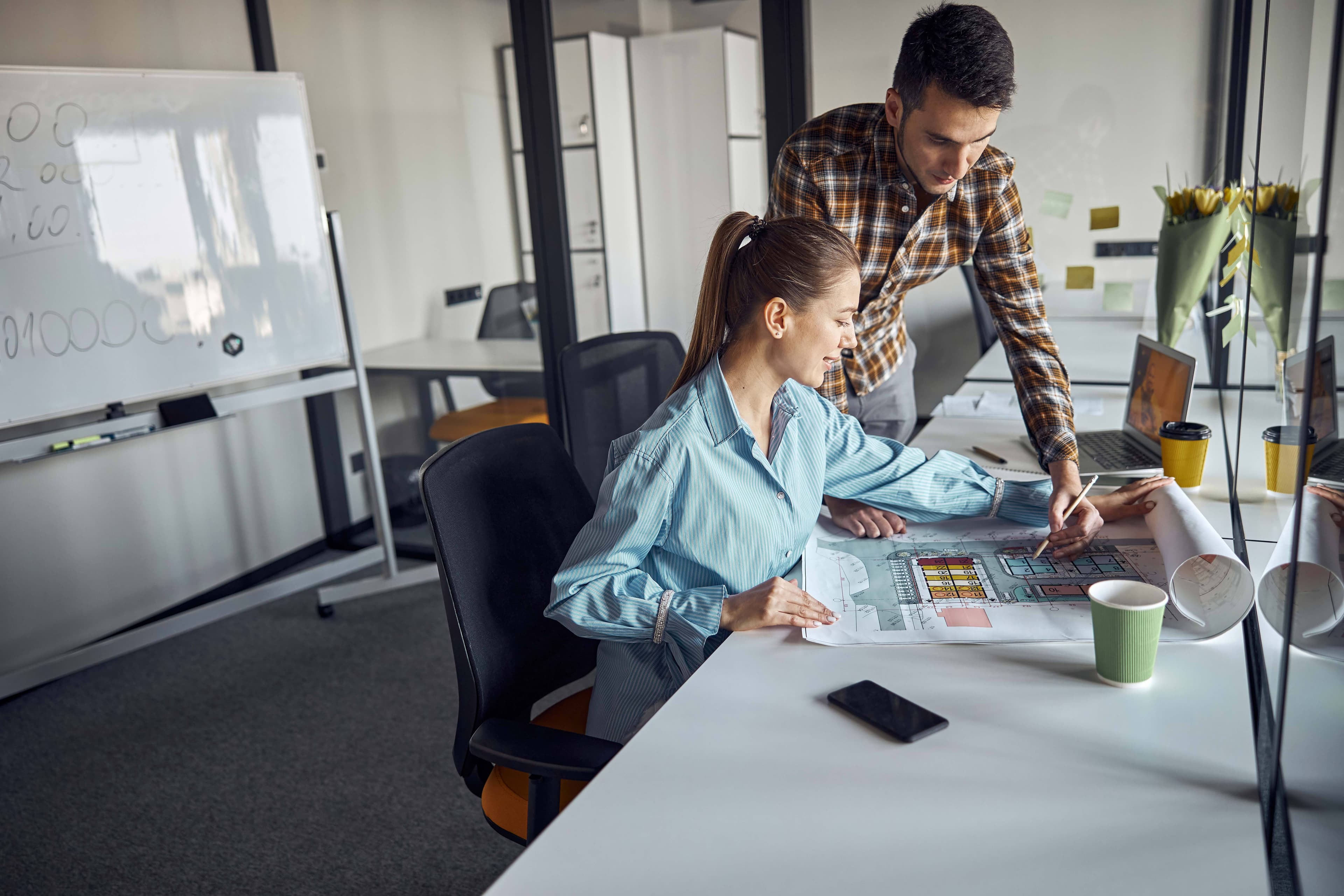 Two professionals collaborating over architectural plans in a modern office, discussing custom glass solutions for shower doors, table tops, and mirrors, with a focus on tempered and beveled glass edges.