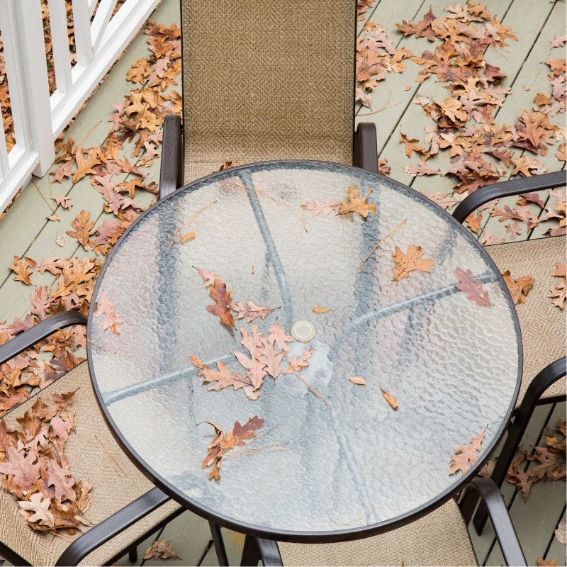 A frosted round glass table and chairs on a deck with leaves on it.