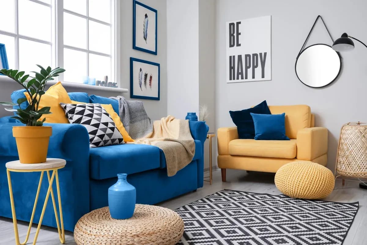 Bright living room featuring a blue sofa with decorative pillows, a yellow armchair, a round mirror, and a geometric rug, showcasing stylish home decor and reflecting custom glass design elements.