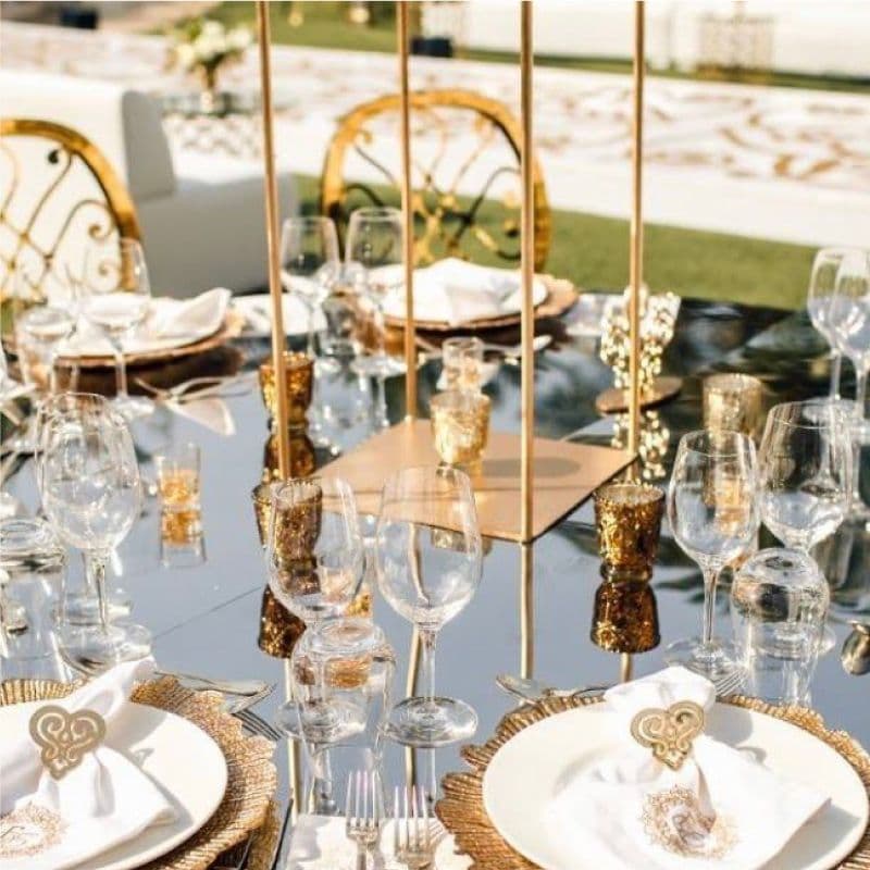 Elegant dining setup on a custom glass table top reflecting golden candle holders and fine tableware, featuring tempered glass elements and an upscale design, suitable for events and celebrations.