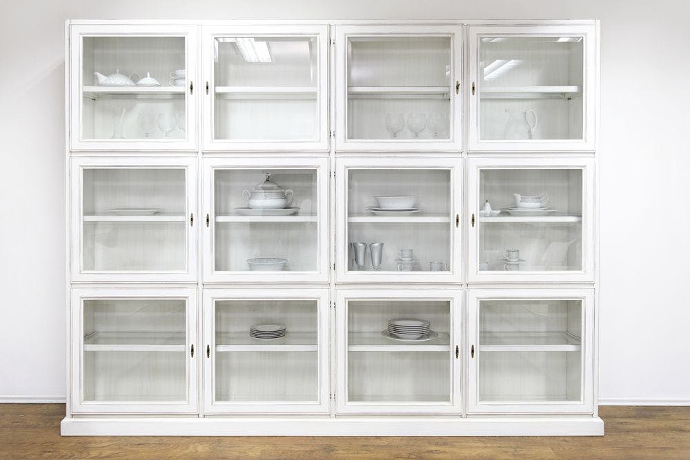 12 evenly spaced square cabinets with glass covers holding items like plates and cups on white wall with brown flooring.