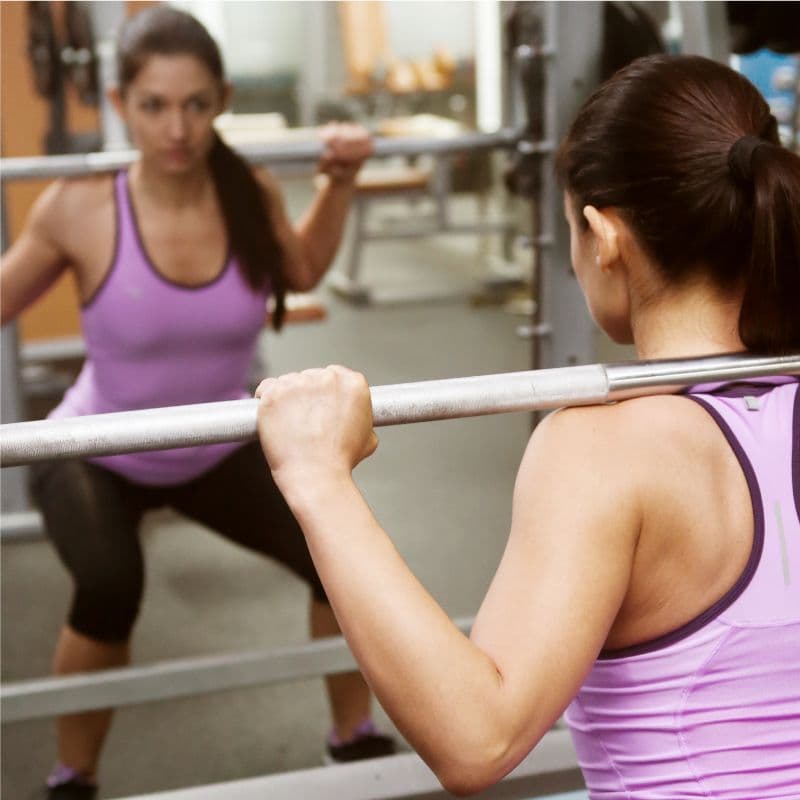 Woman lifting weights in a gym, showcasing strength training with a reflection in a mirror, emphasizing fitness and wellness.