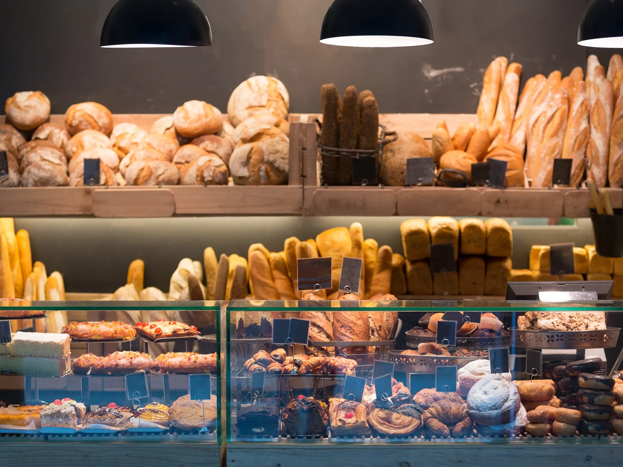 A bakery's clear acrylic display case filled with fresh pastries, with shelves of bread and baguettes in the background.