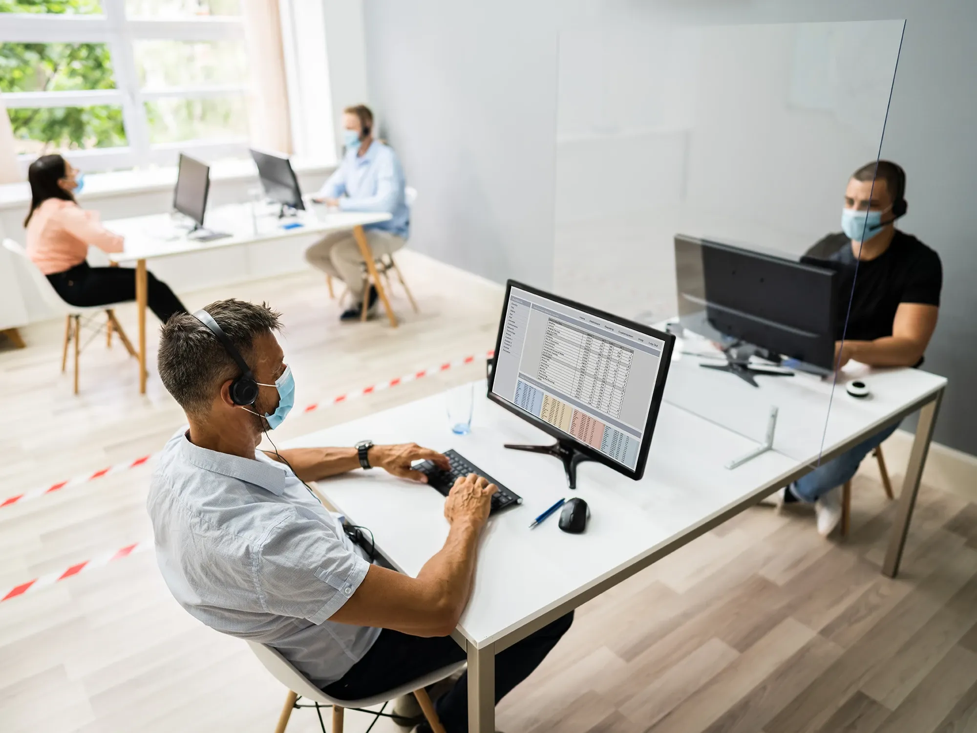Office workers at socially distanced desks with a clear acrylic plexiglass sneeze guard separating them for safety.