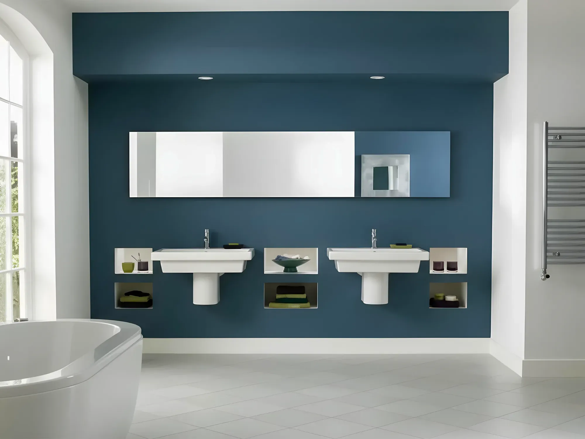 A modern bathroom with a bold teal accent wall, two white pedestal sinks, and a long, horizontal custom-cut mirror.