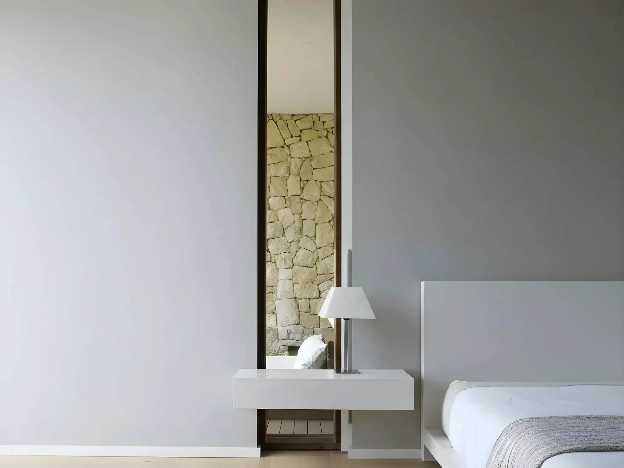 A minimalist bedroom featuring a tall, narrow, custom-cut mirror built into a wall niche, reflecting a stone accent wall.
