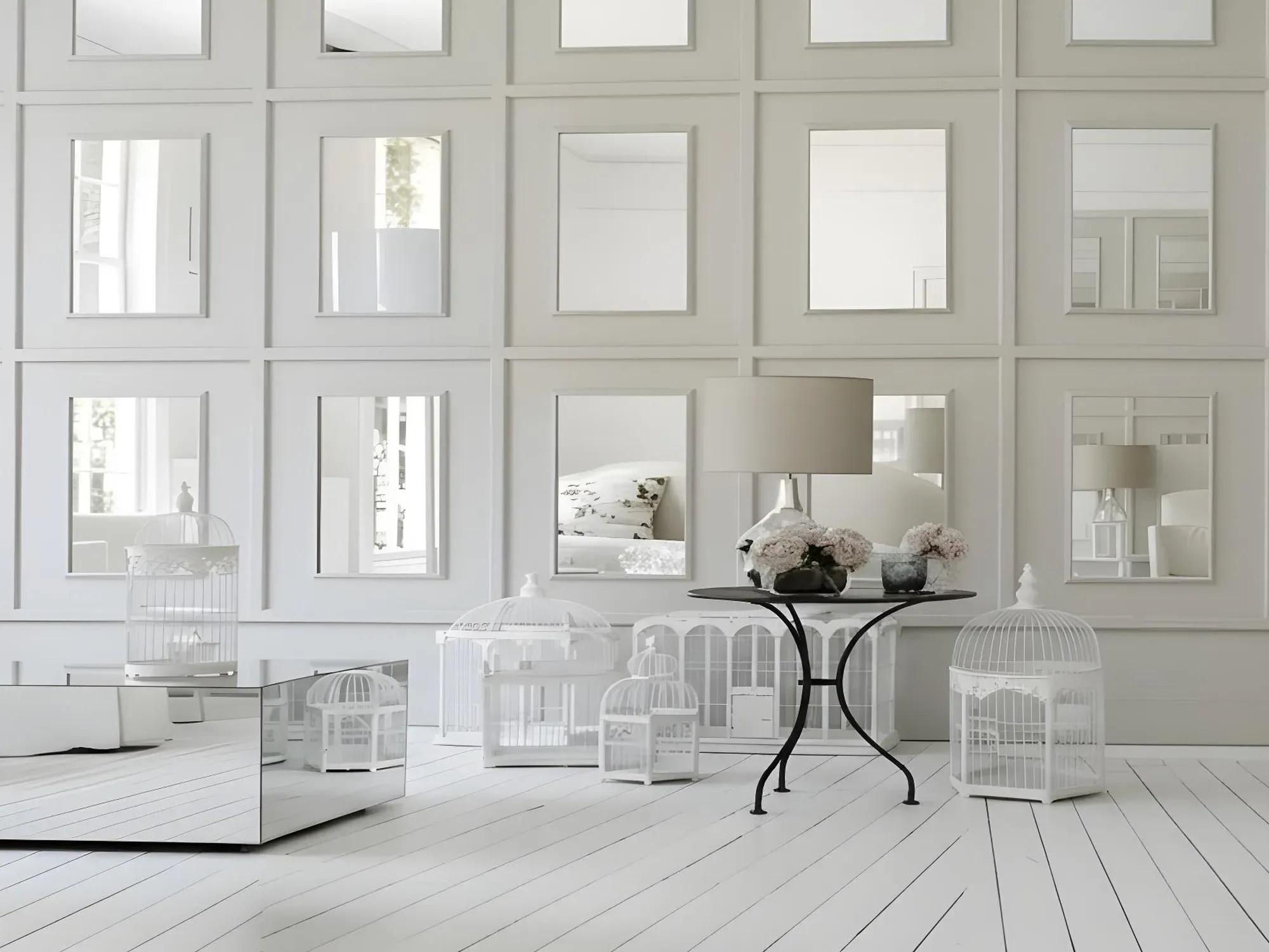 A whimsical all-white room with a wall of custom-cut rectangular mirrors inset into decorative paneling, surrounded by white birdcages.