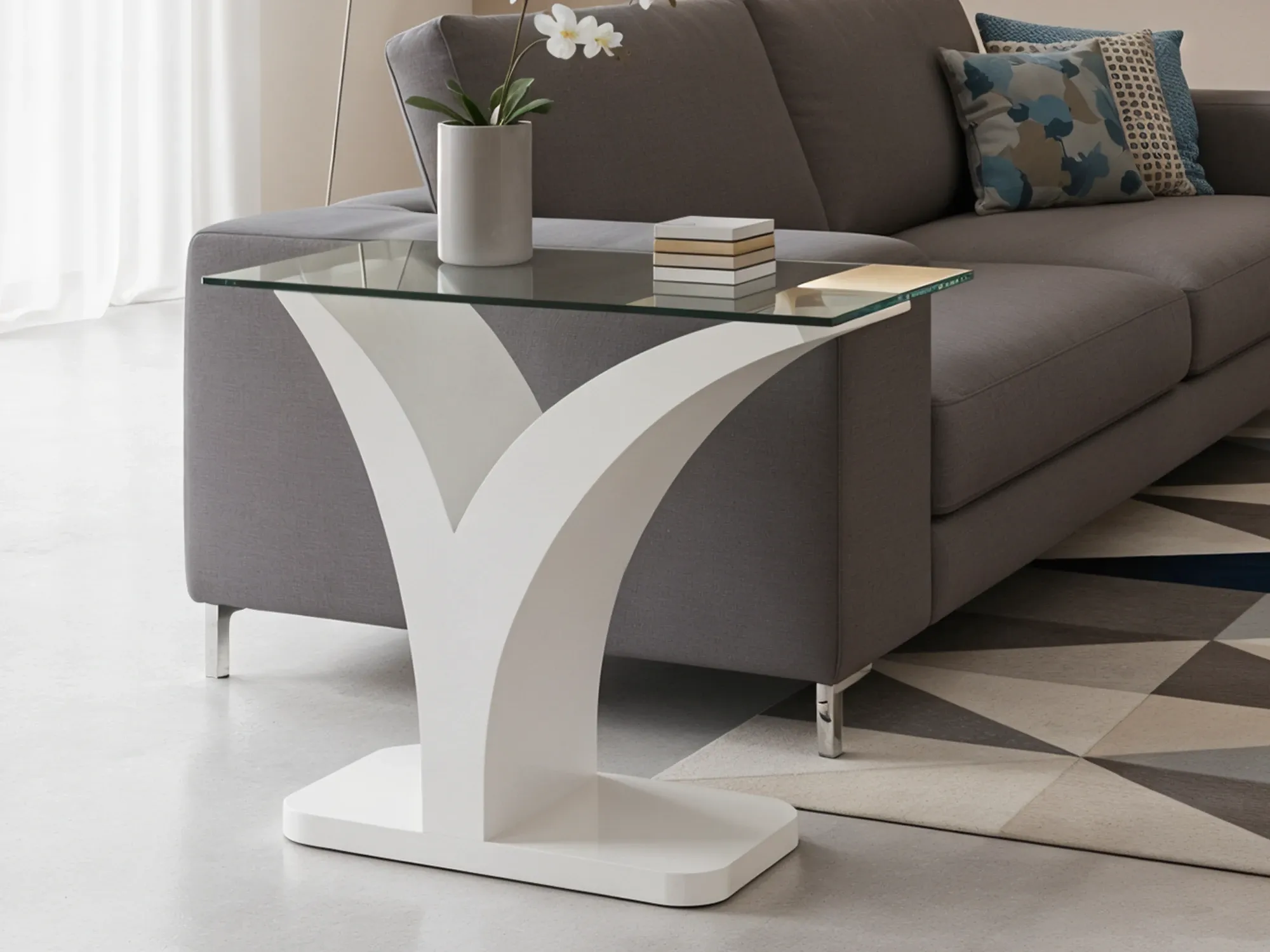 A modern glass-top side table with an elegant, white Y-shaped pedestal base, placed next to a grey sofa on a geometric rug.