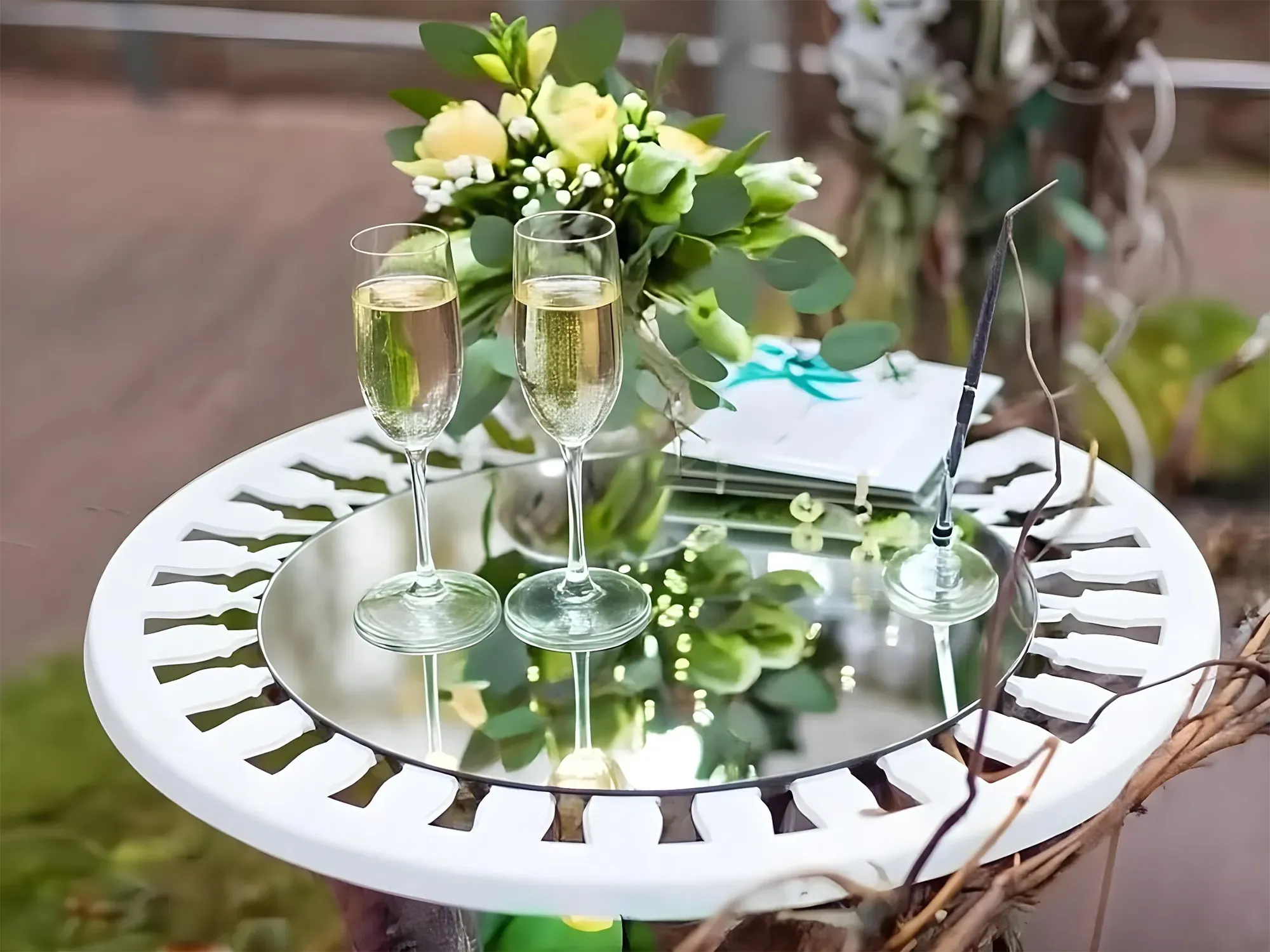 A close-up of a white wedding table with a round mirror top, reflecting two glasses of champagne and a floral bouquet.